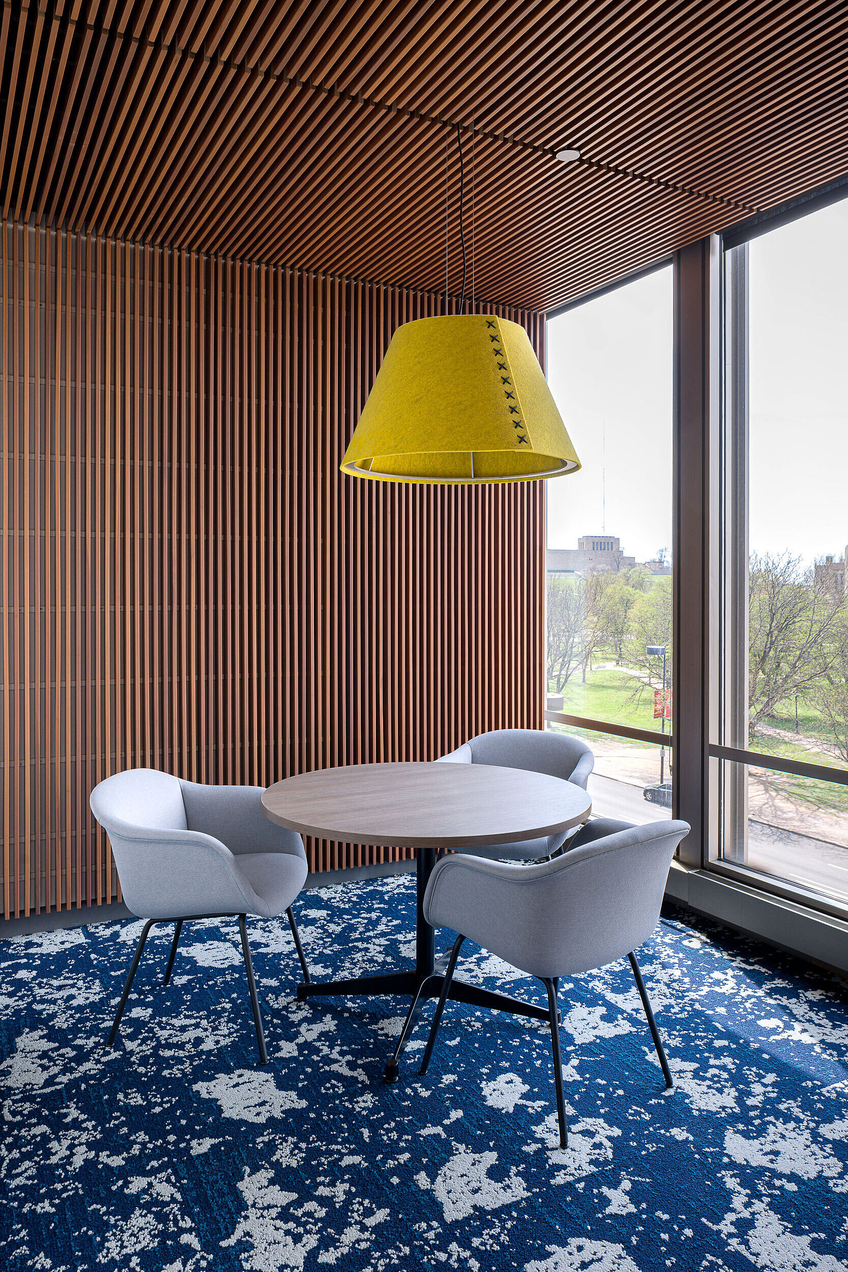 Nook with slatted wall & ceiling has a round table and 3 blue upholstered chairs. A dramatic yellow lightshade hangs above. Dark blue/white graphic carpet.