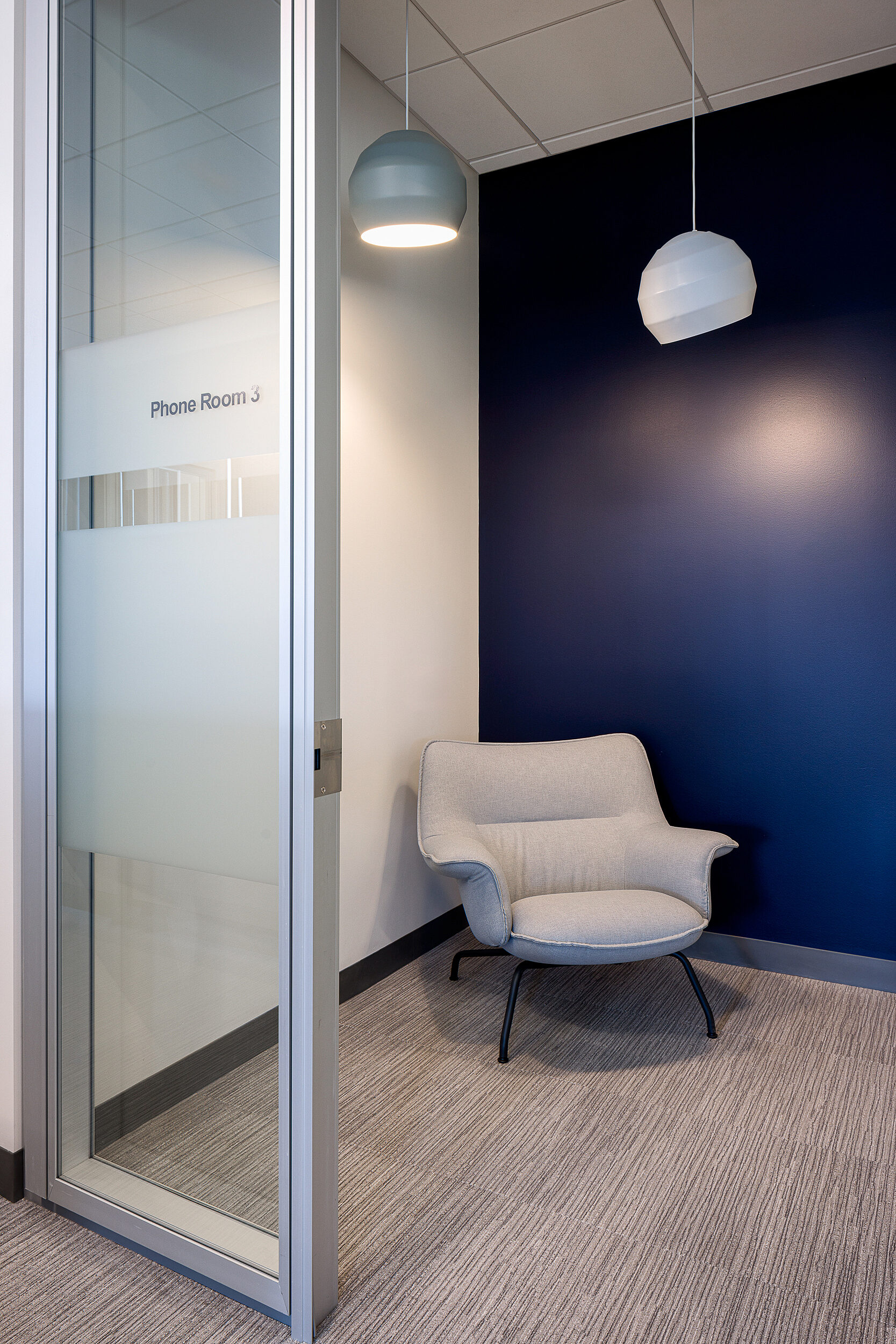 Employee phone room with a grey upholstered lounge chair with arms and black legs. Dark blue accent wall and two hanging lights in grey and white above.