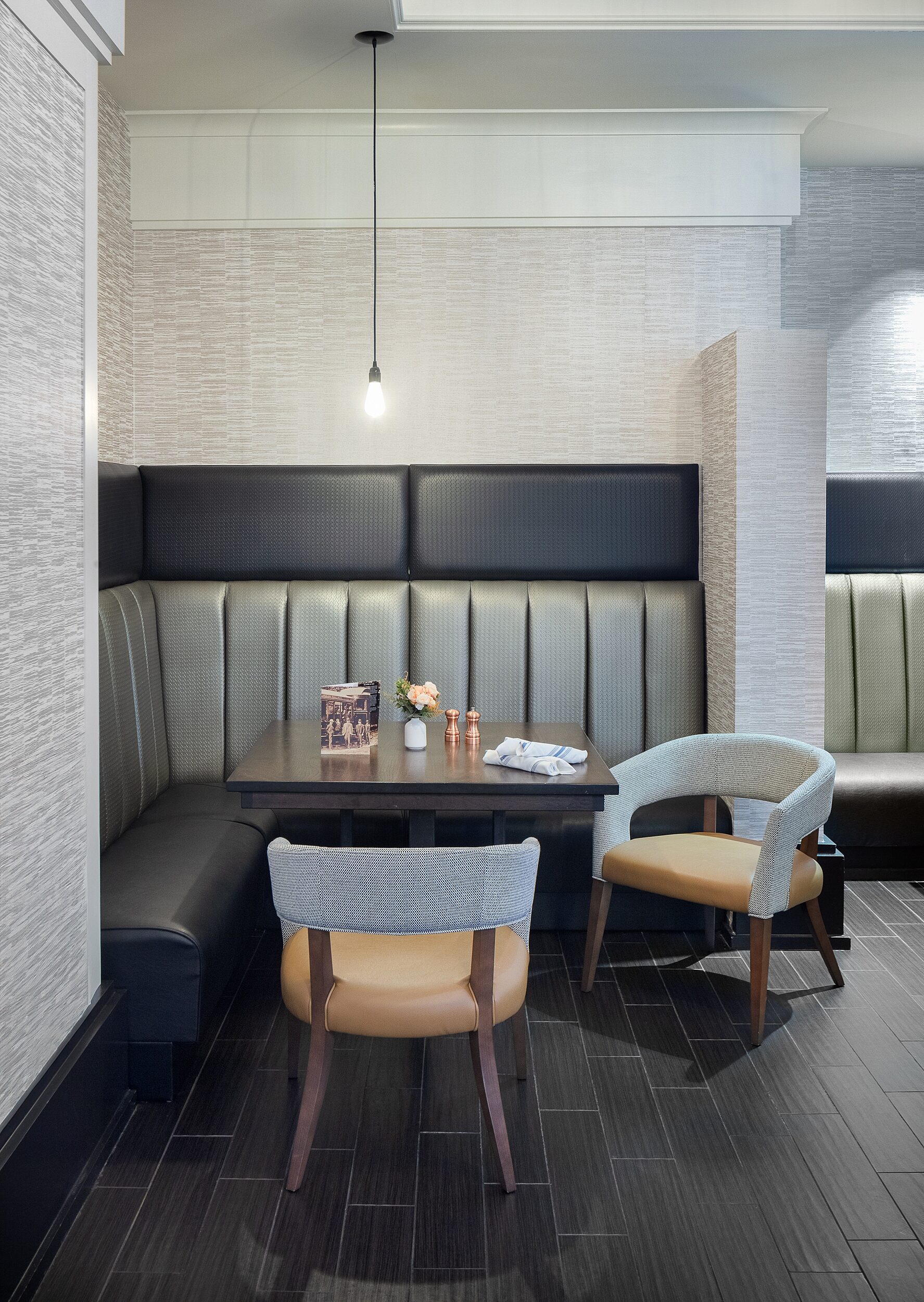 Sheraton Overland Park corner booth with Silver/grey banquette seating with a cafe table and grey and brown chairs. Single lightbul lamp hangs from ceiling