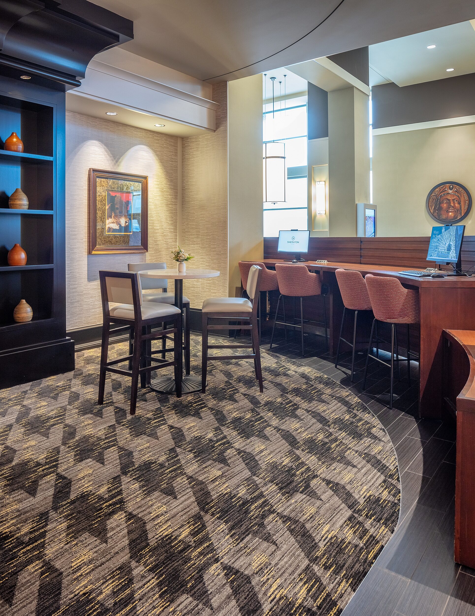 Hotel business center with computer area. Red patterned tall stools. A tall cafe table with white top and dark wood backed stools with white leather