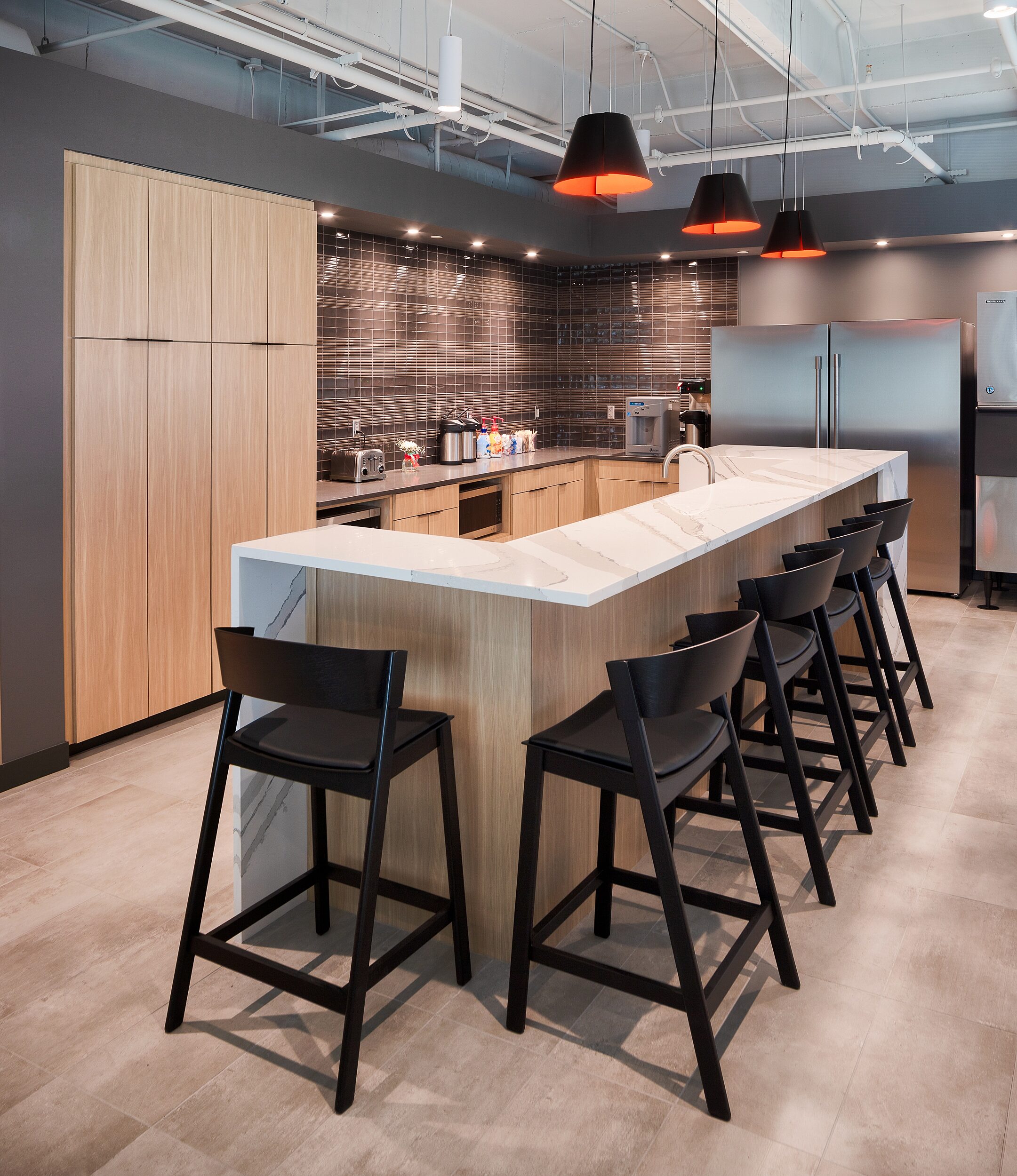DRI - breakroom kitchen with marble counter and black bastools with leather seat cushions. Three black drop lights hang over bar with red insides.