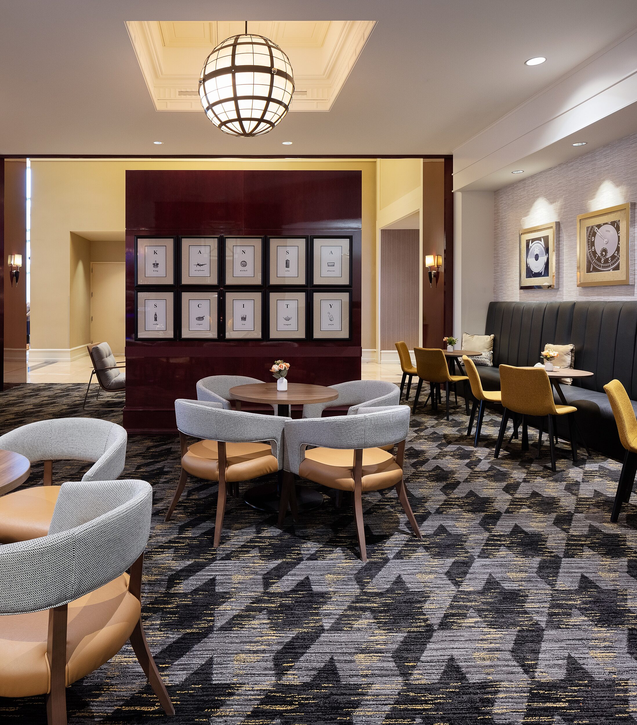 Hotel lounge. Wood cafe tables and upholstered grey/brown cafe tables. Banquette seating on right. Partial wall with artwork that spells Kansas City.