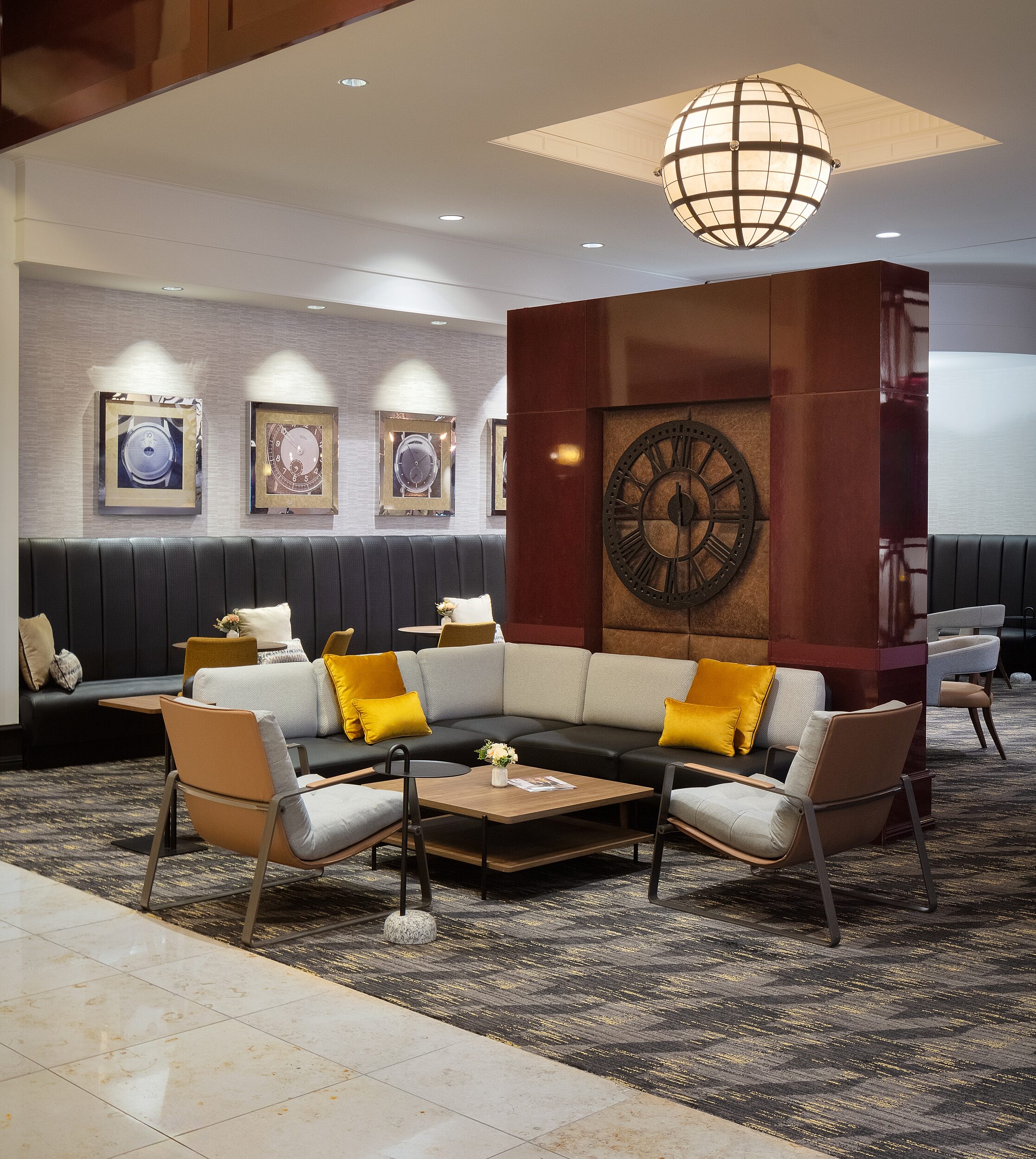 Hotel lounge L-shaped leather sofa with pillows. Two lounge chairs and two-tiered wooden coffee table. Large clock on accent partial wall. Globe light.
