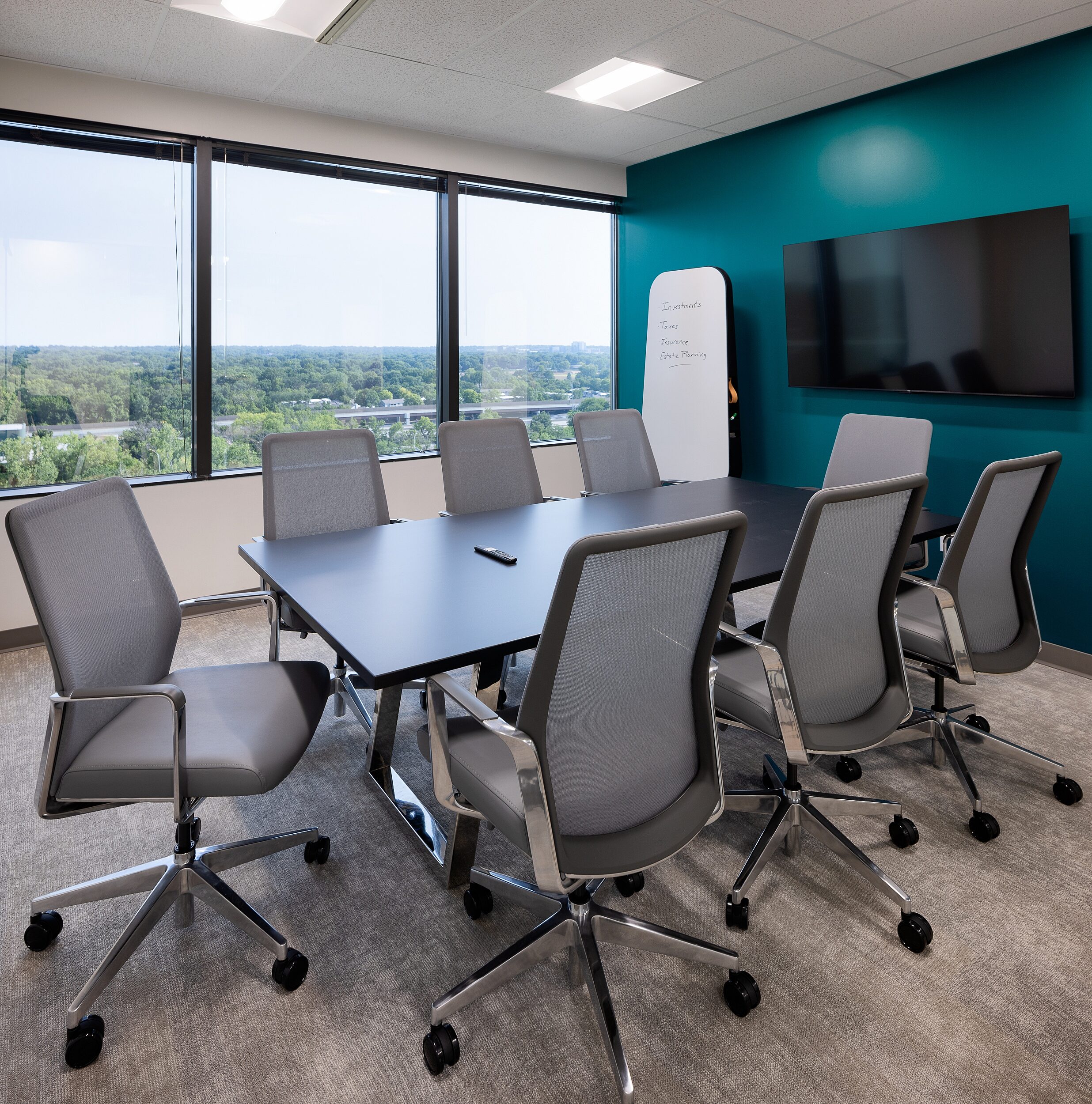 Conference room w/ teal accent wall and treed view. Black/chrome conference table with 8 high-back rolling chairs with grey upholstered seat/chrome base