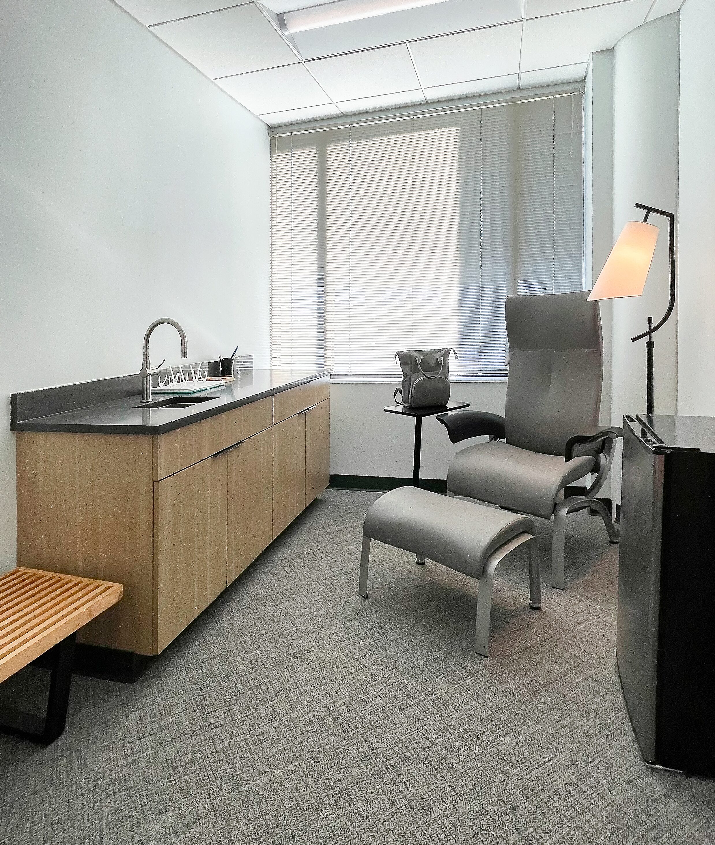 DRI Mother's Room - Room with Sink, a small fridge, a Herman Miller Bench and a grey Nemschoff chair and ottoman. A small lamp sits next to the chair.