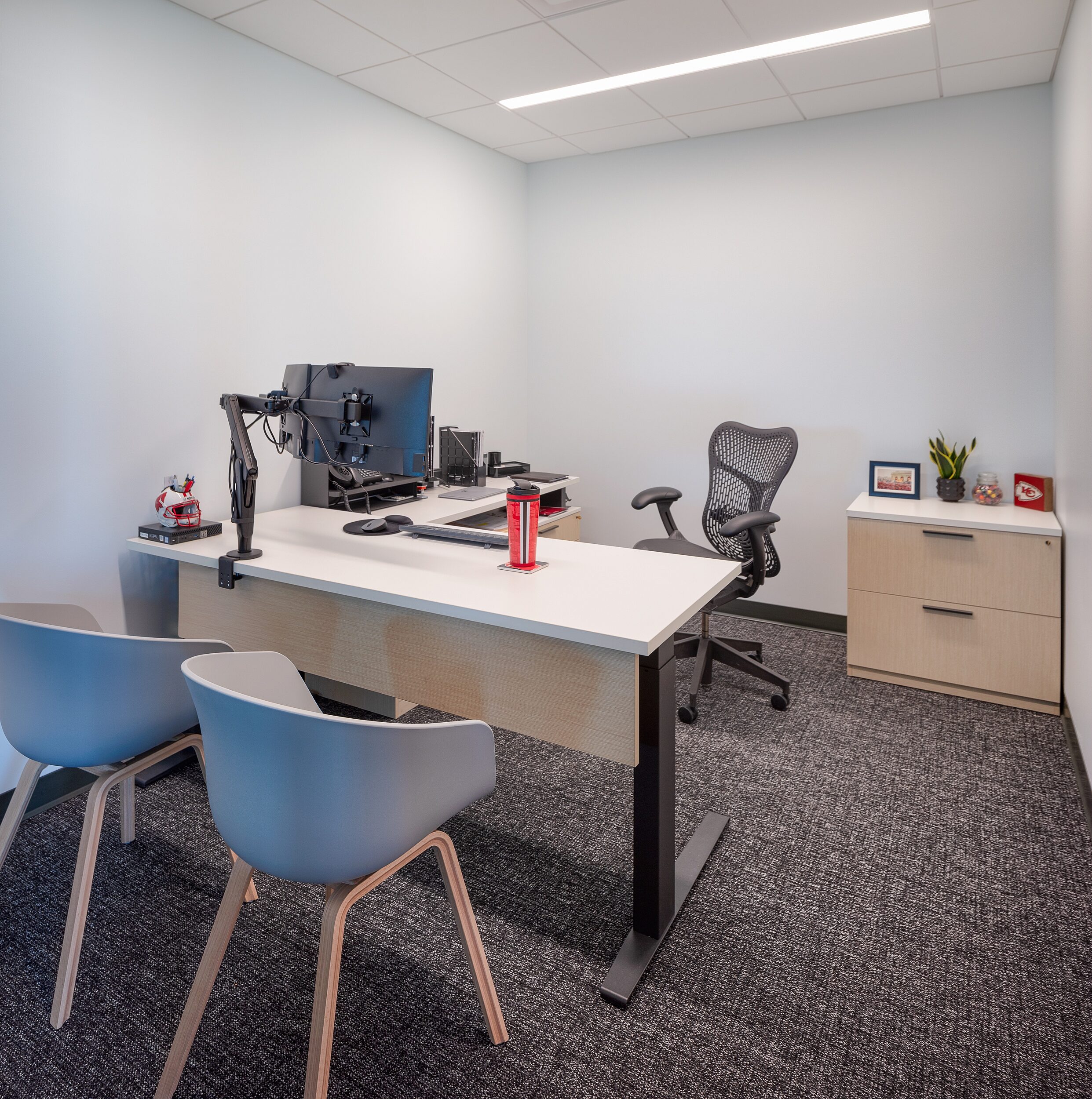 DRI private office with height adjustable L-shaped desk, ergonomic chair, two grey guest chairs with wooden legs and small storage cabinet in back corner