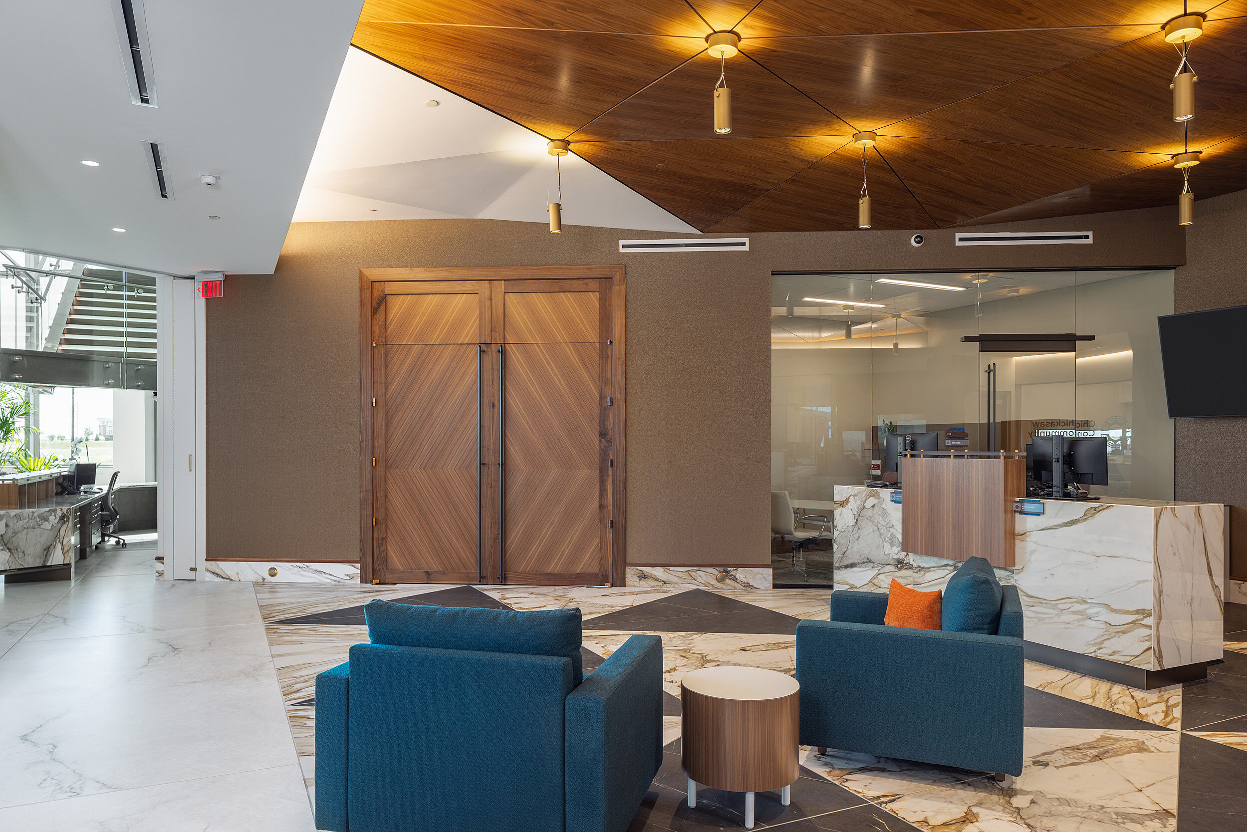 Bank lobby/reception area with a pair of blue lounge chairs and a drum-shaped side table. Marble floor/reception desk, wood accents doors and ceiling.