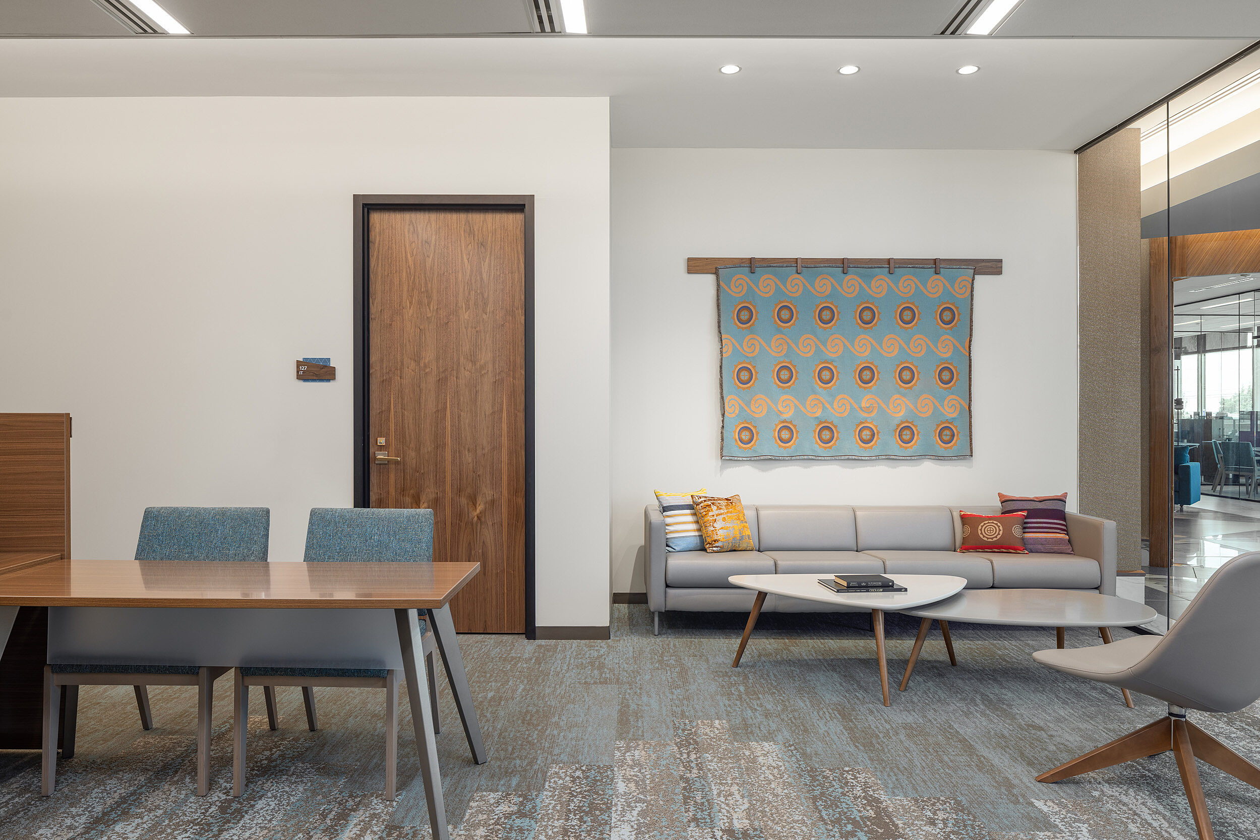 Collaboration area outside of private offices with table, grey leather sofa and chair, triangular nested coffee tables, and a tapestry on the wall.
