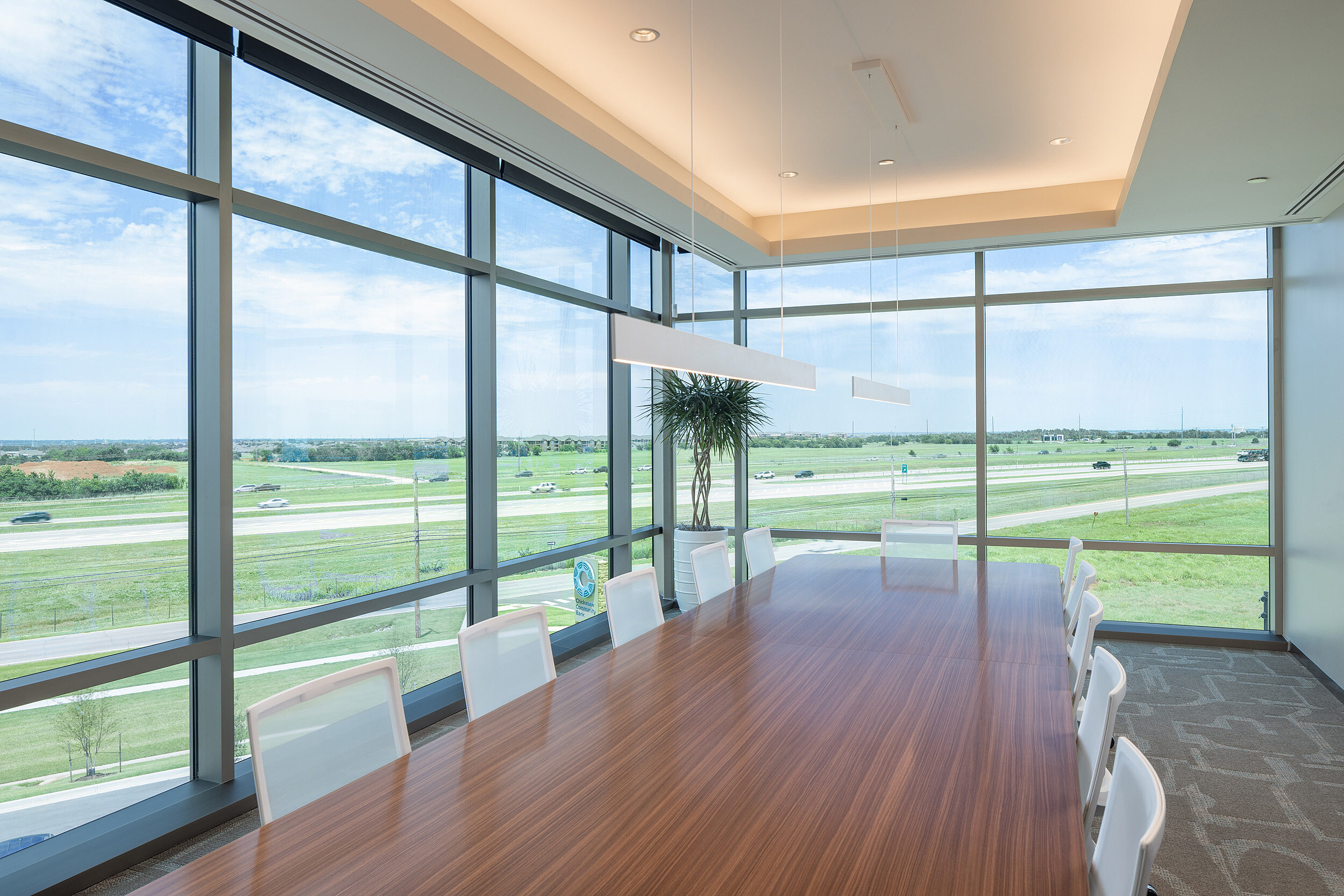Large conference room with wooden table and white chairs. Two walls are floor to ceiling glass with a view of the highway.
