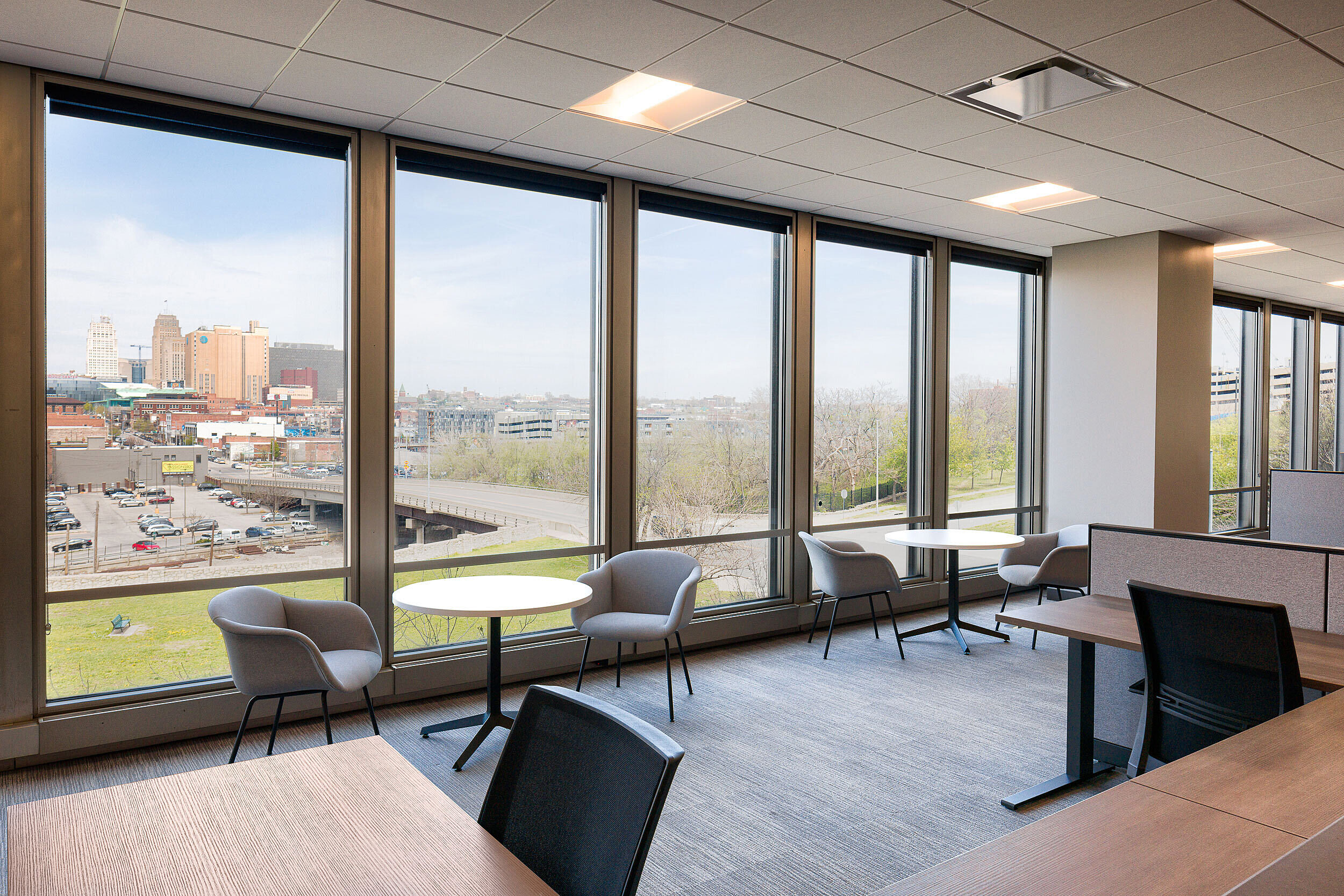 Open office area with back-to-back workstations. Two Cafe tables with two blue upholstered chairs are in front of window with view of downtown Kansas City.