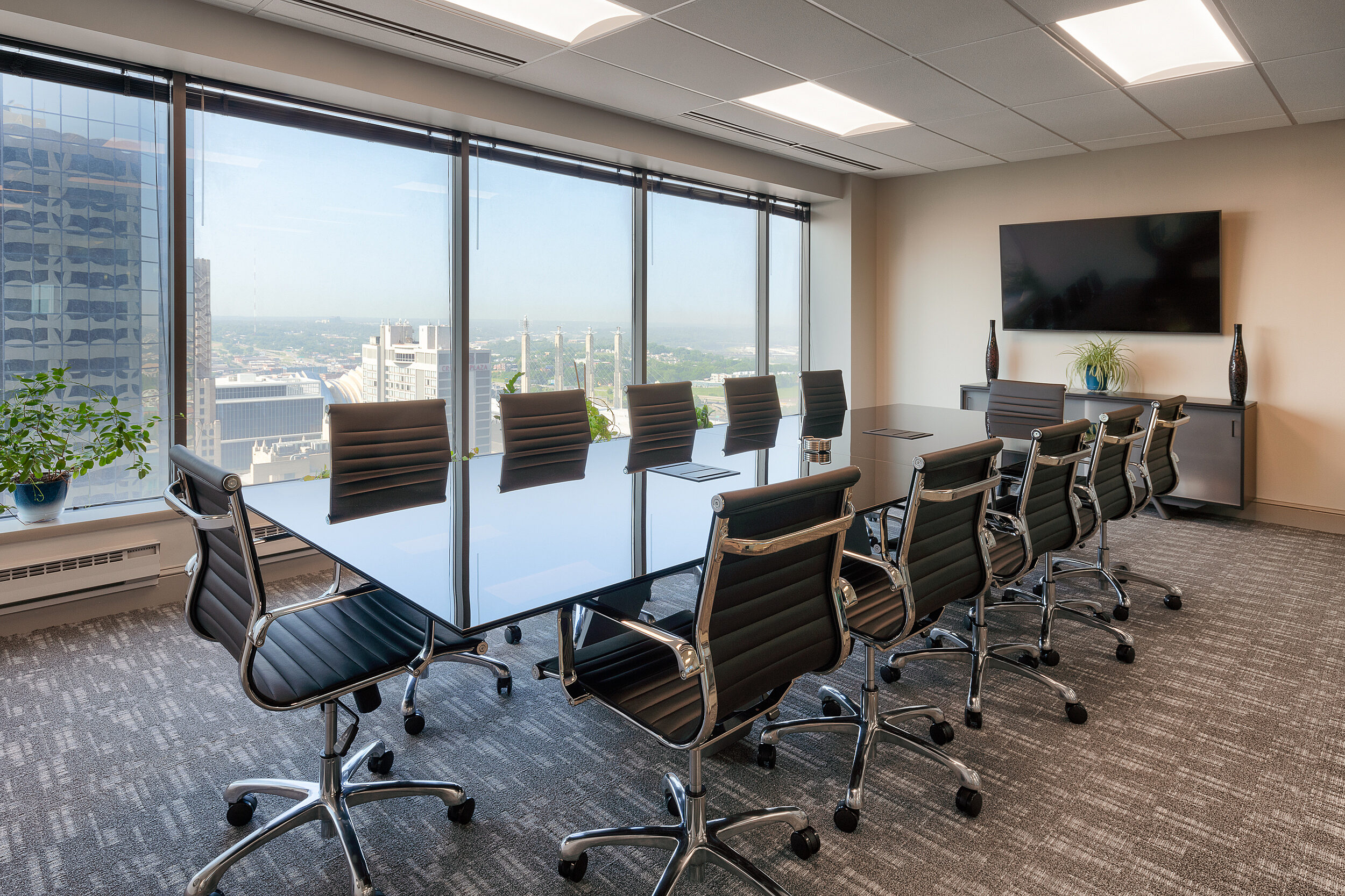 Conference room with skyline view. Large table surrounded by 12 black Eames aluminum chairs. Credenza at end with mounted television above.