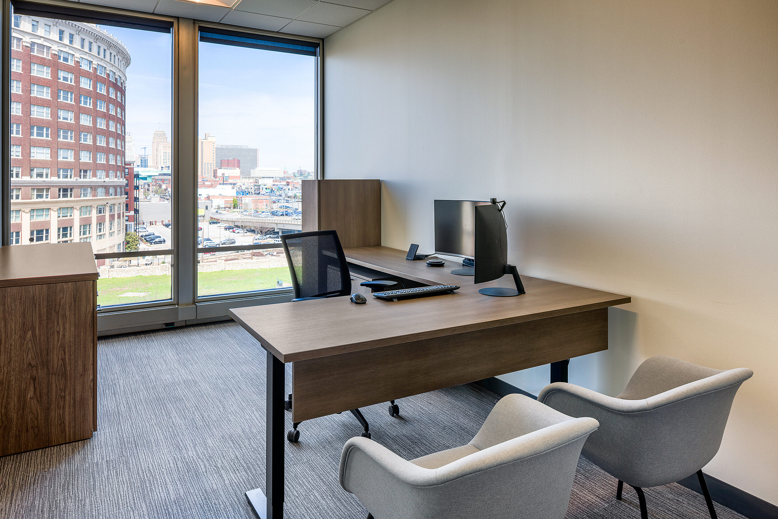 Private office with L-shaped height adjustable desk w/ locker and storage unit. Two blue guest chairs. Kansas City's former Western Auto building in window