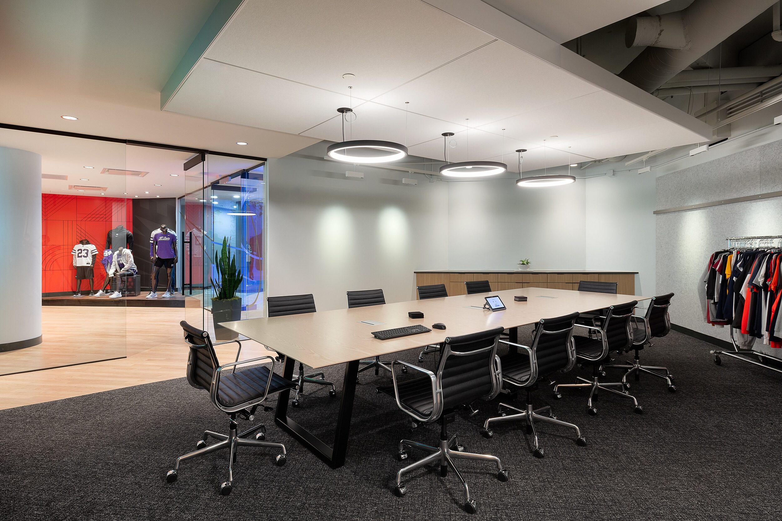 DRI Executive Conference Room with black Eames Aluminum chairs. Mannequins wearing sportwear seen in adjoining room and rack of sports apparel on back wall