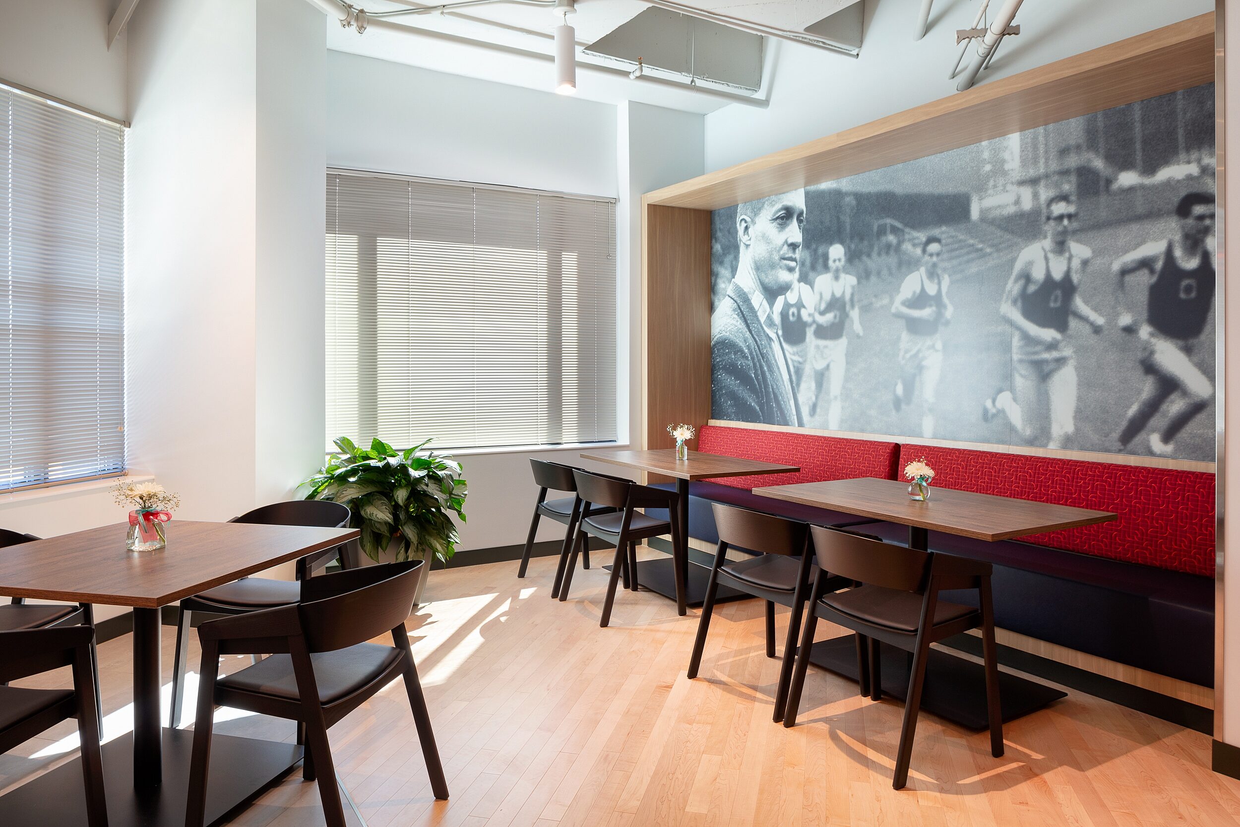 DRI breakroom seating with red/orange quilt fabric, two tables and black/white mural wall with athletes running. Four top table with leather seat cushion