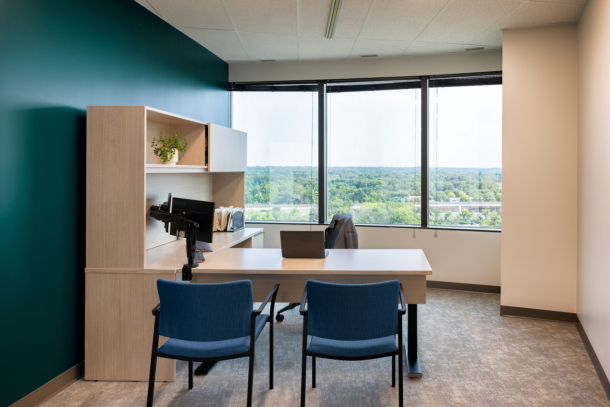 Private Office with teal accent wall, light wood desk with two overhead cabinets, two dark blue side chairs, windows show view of trees and a highway