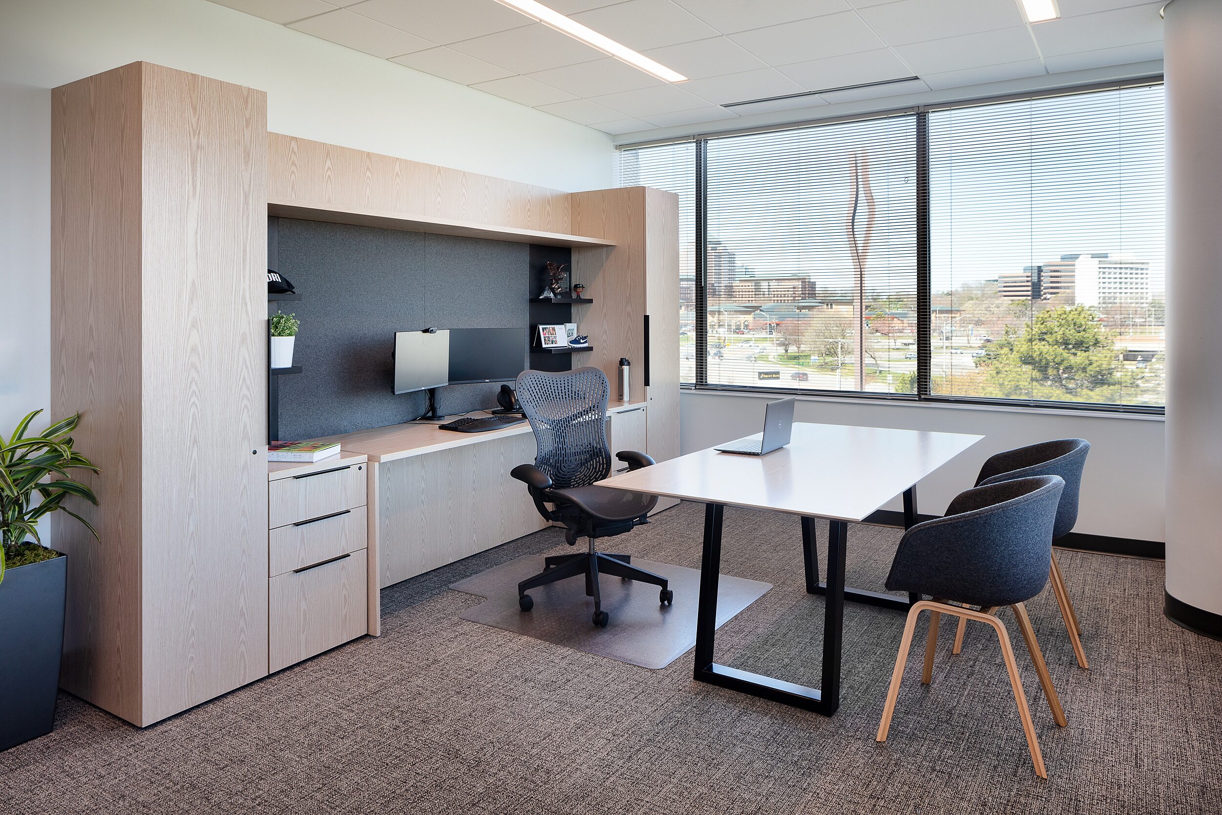 DR Executive Office with large desk with wall unit in light wood and separate desk/table with two u-shaped guest chairs. City view in large windows.