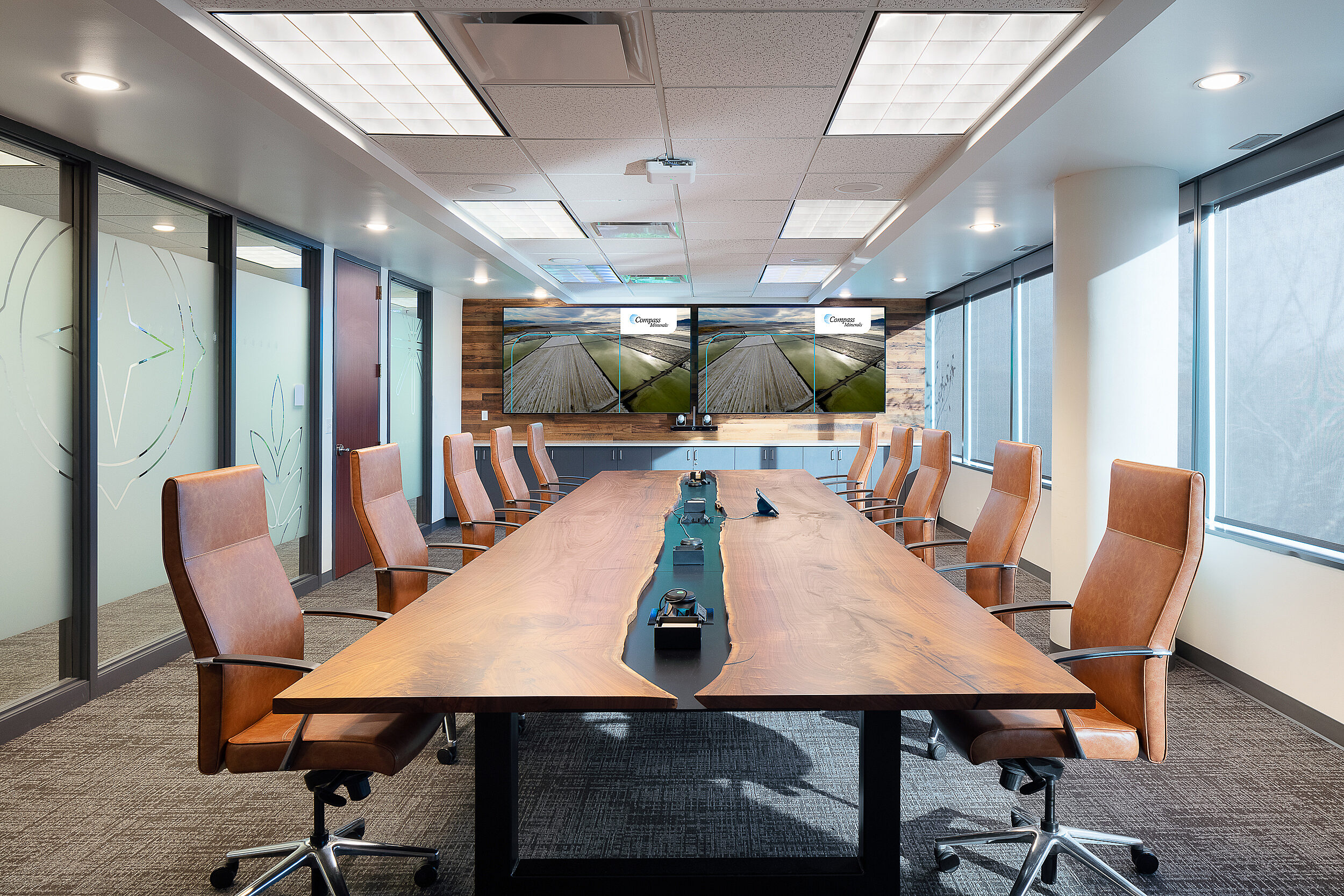 Compass Minerals boardroom with frosted blass wall. Custom table with live edge island (exposed black) along width and sleek, high-backed leather chairs.