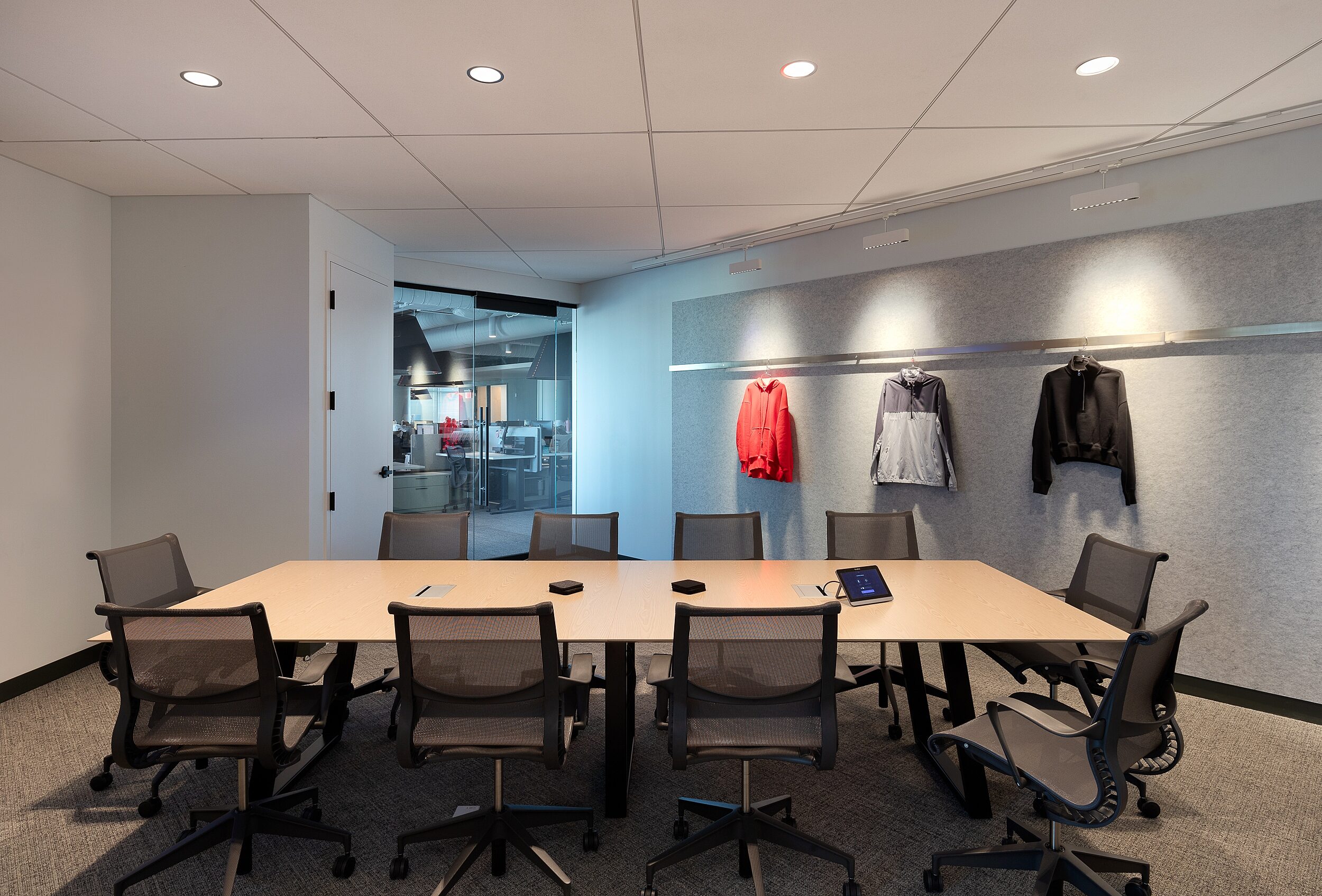 DRI Larger conference roo with metal rail to hang sports apparel. Sliding glass doors open into employee area filled with multiple workstations.