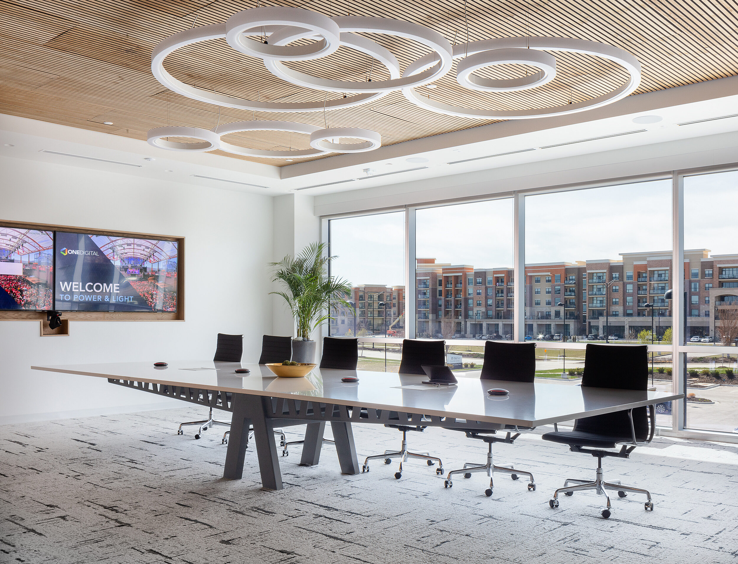OneDigital executive conference room with Halcon table with sawhorse base and black chairs. Circular lighting and wood slat ceiling. Two large TV screens.