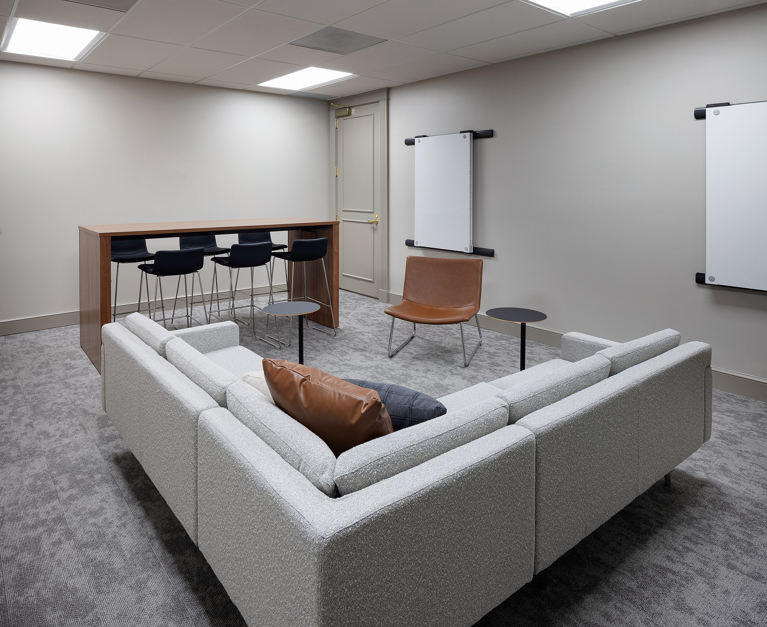 Collaboration room with grey, upholstered L-shaped sofa, tall counter-height table with black stools. Black and brown pillows on couch, Brown shell chair