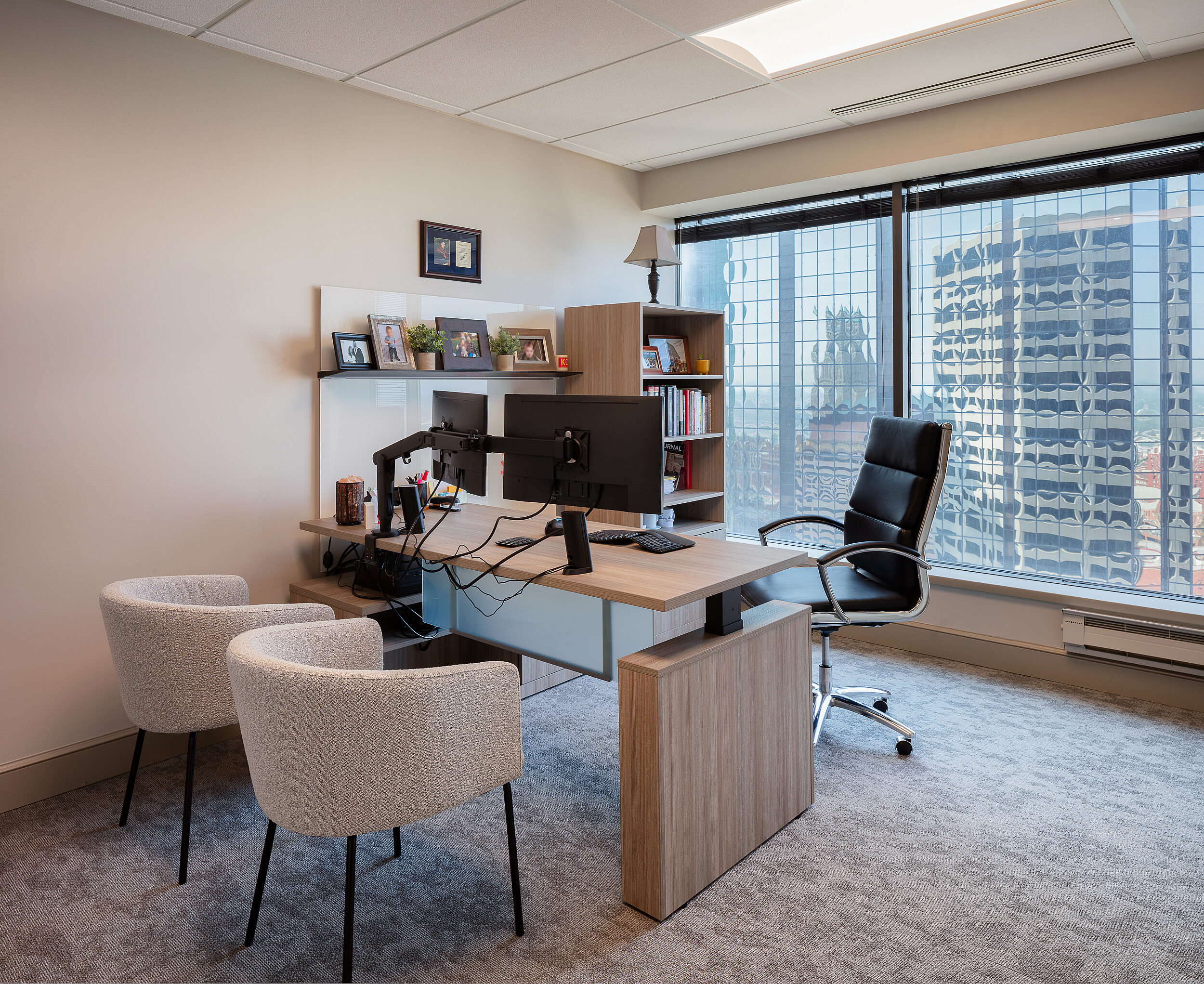 Law firm office with pale wood, height-adjustable desk and rolling task chair. City building in big window. Upholstered, curved back guest chairs in front