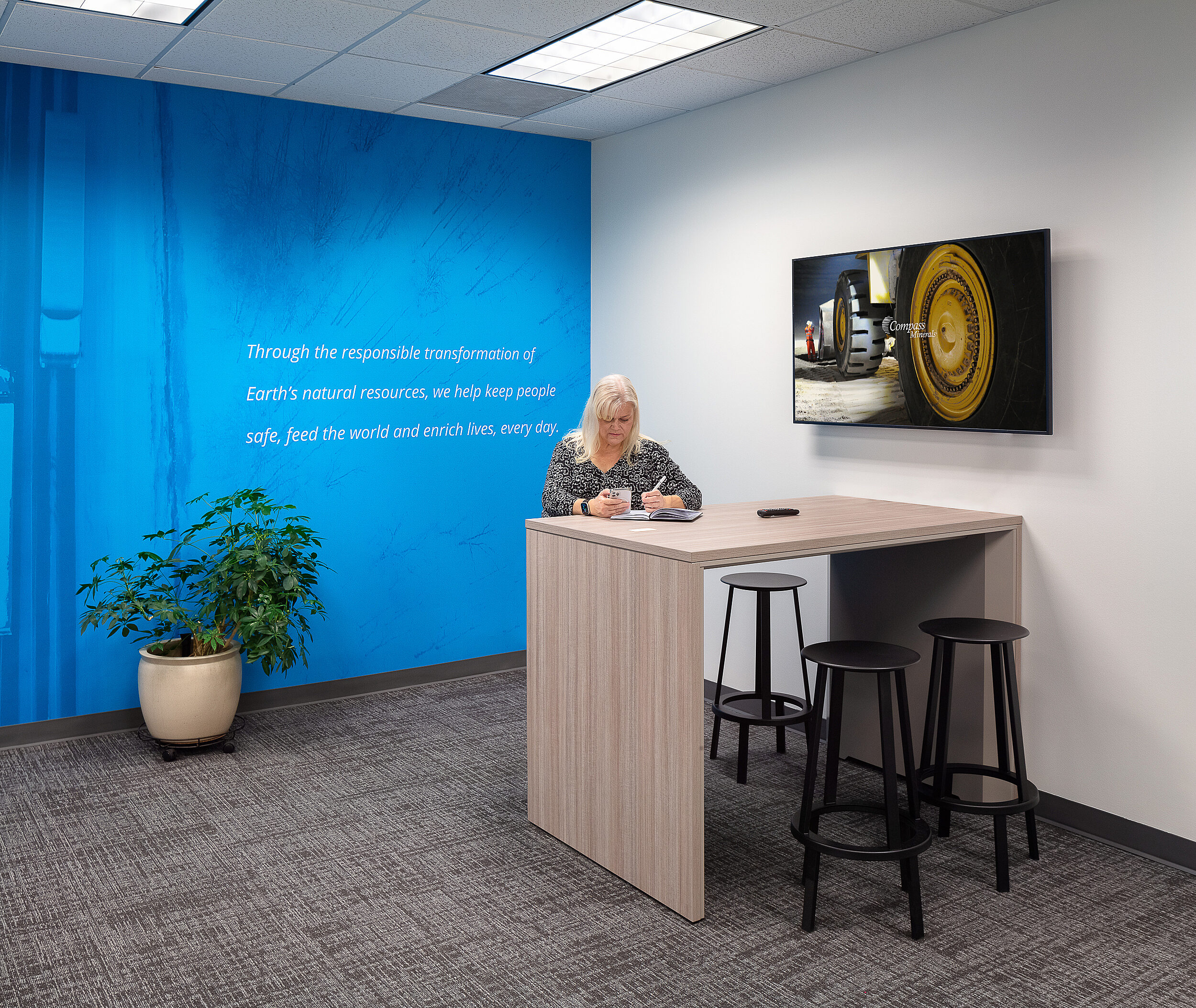 Compass Minerals collaboration area with work counter and stools and blue accent wall.  Woman sits on stool on phone call taking notes.
