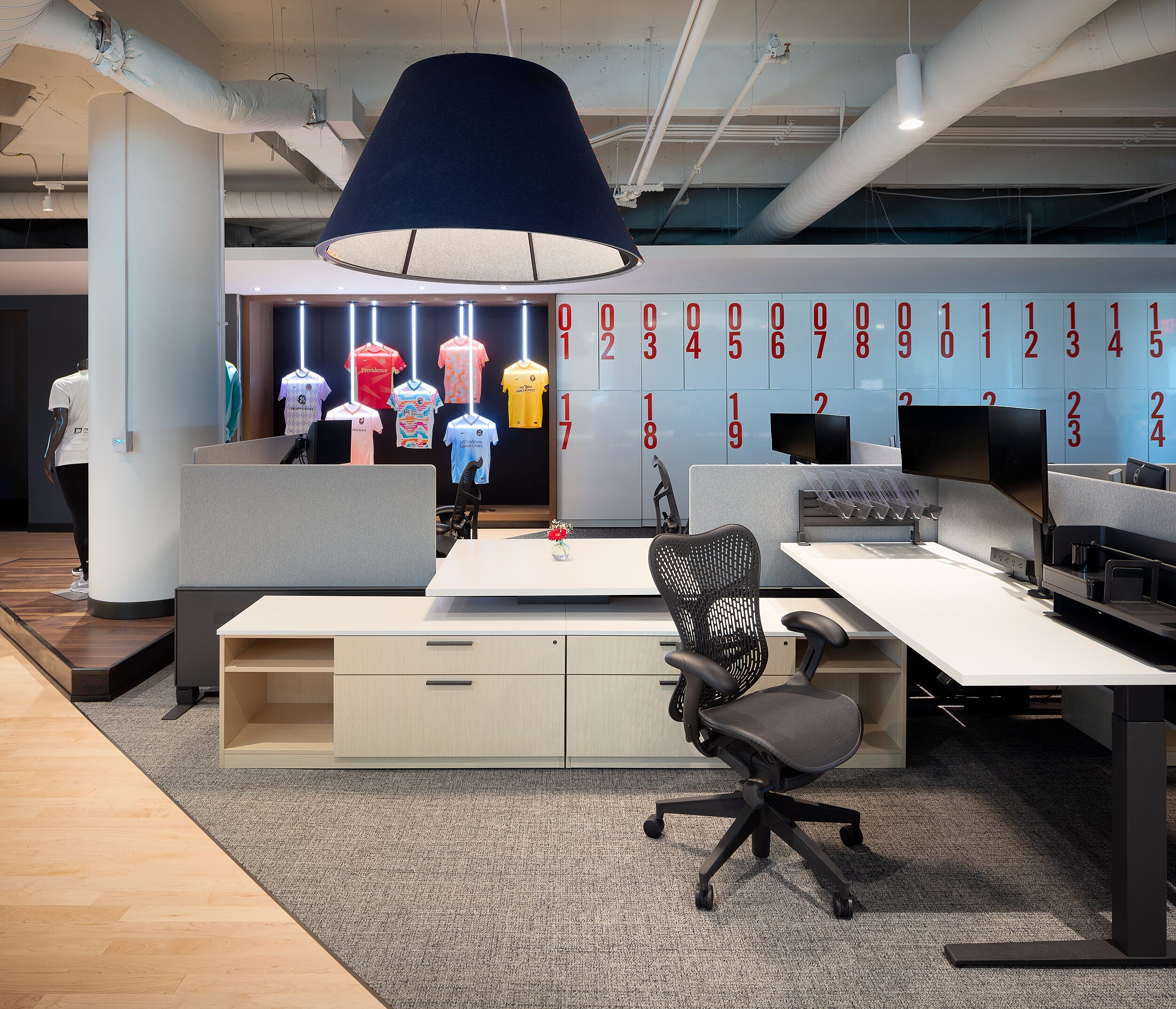 DRI Workstation 4-pack. Apparel display hangs in background with locker all with large red numbers. Height-adjustable desk with dual monitor and storage.