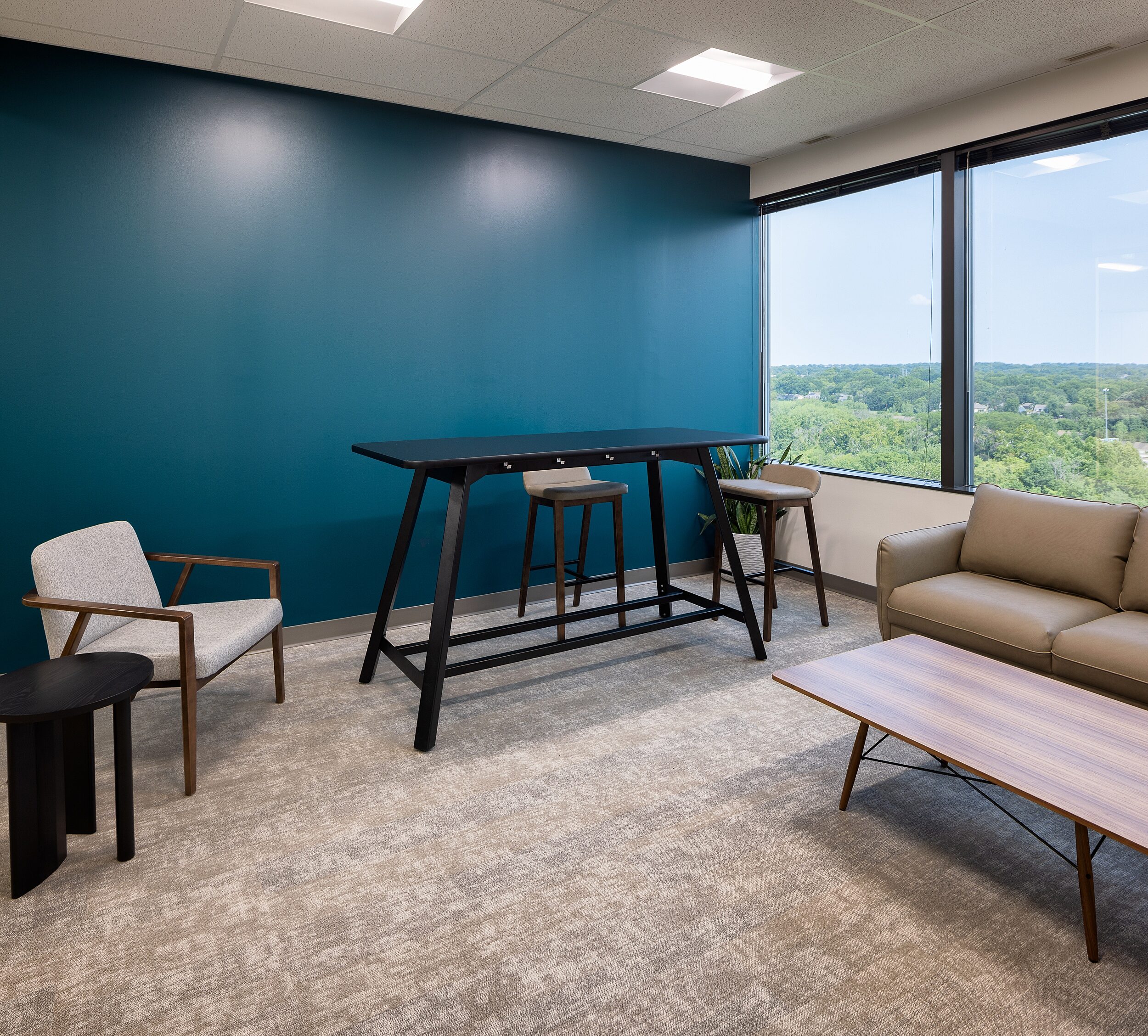 Collaborative room with dark teal wall, tall collaborate table with stools, a beige leather sofa and coffee table and 2 lounge chairs with rich wood detail