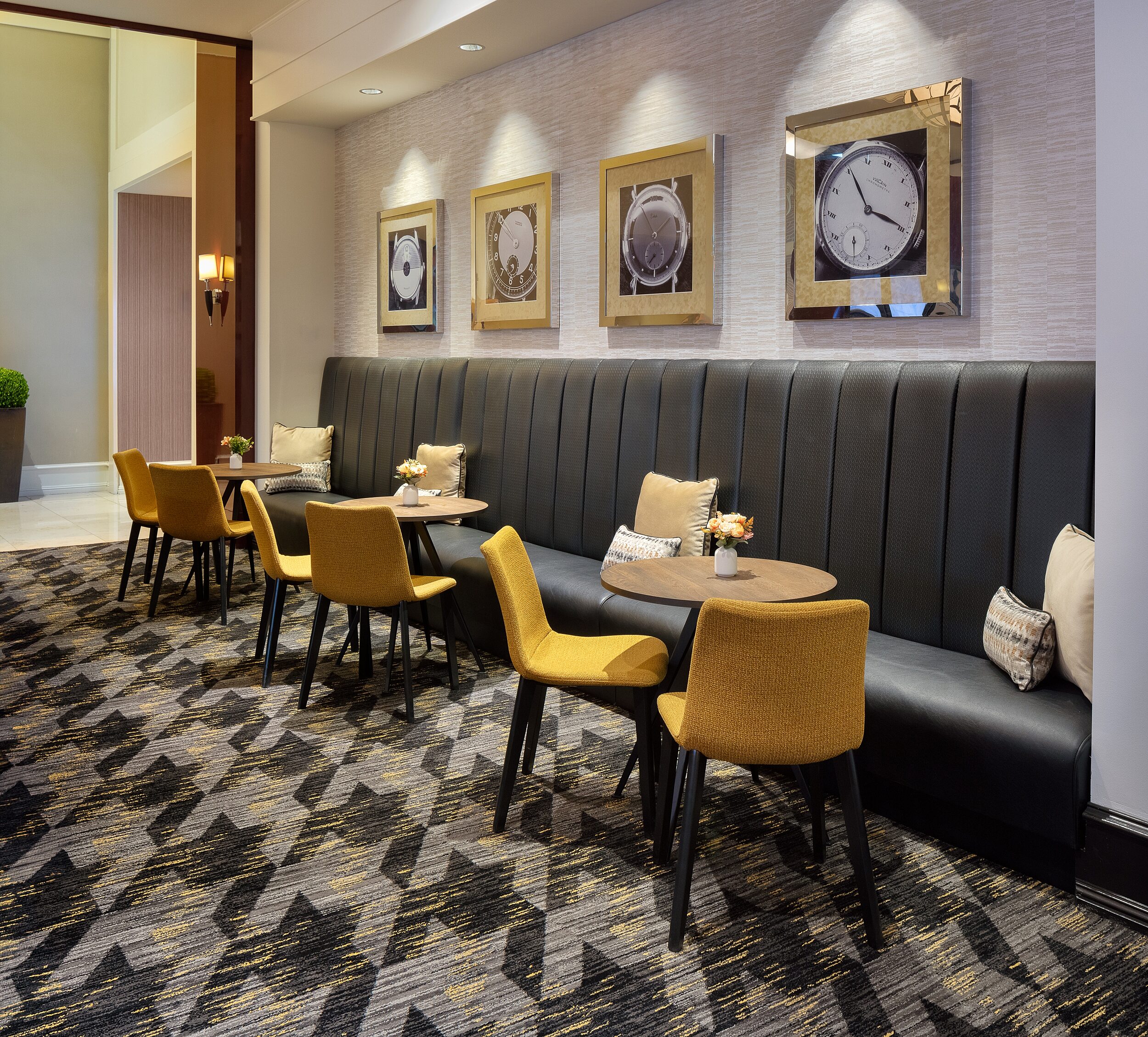 Hotel lounge area with banquette seating along wall with throw pillows. Small wooden cafe tables and yellow upholstered chairs. Bold patterned carpet.
