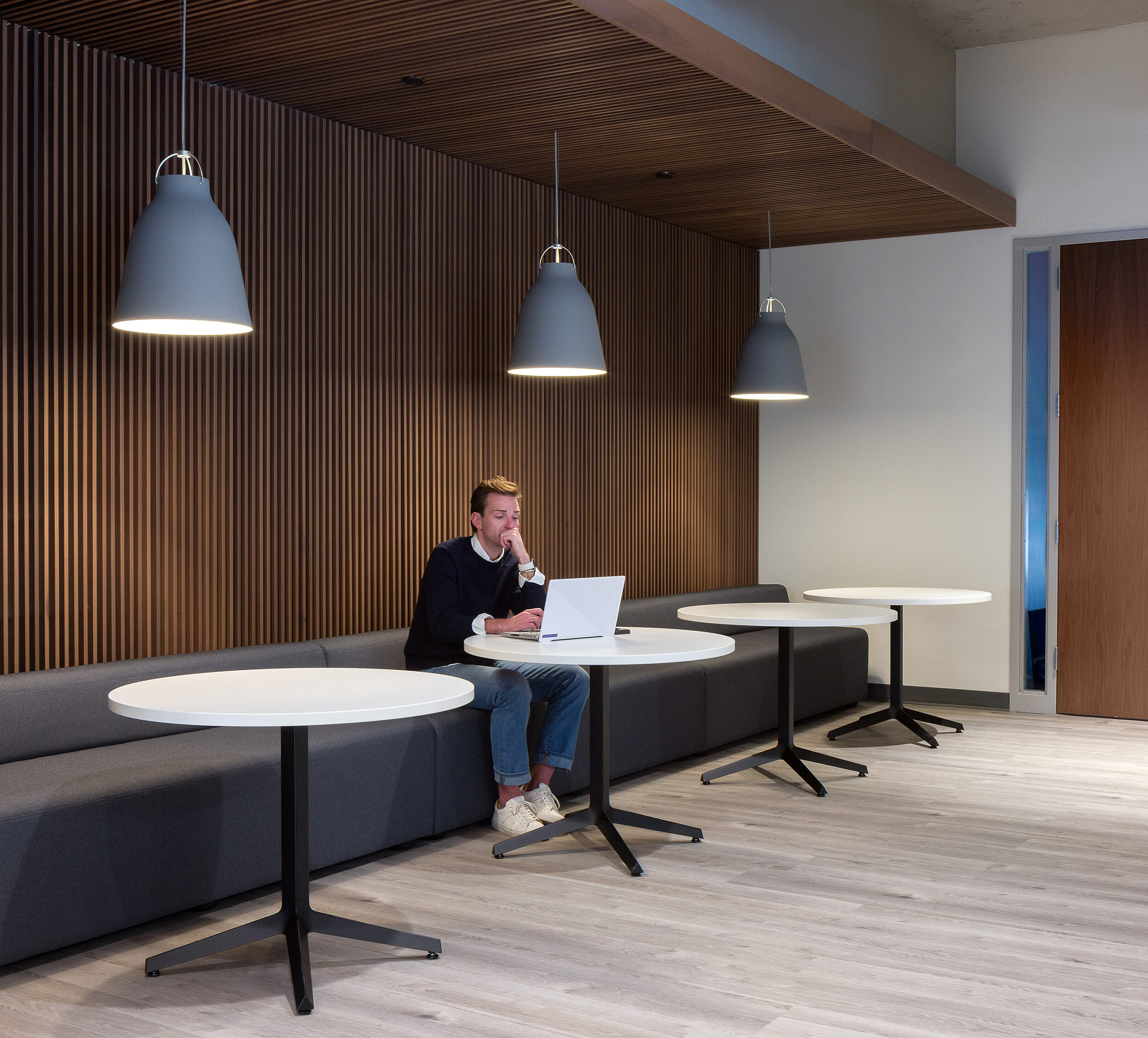 A man works on laptop. Sits in a nook with grey banquette seating with round cafe tables with grey pendant lamps above them.  Slatted wood wall & ceiling.