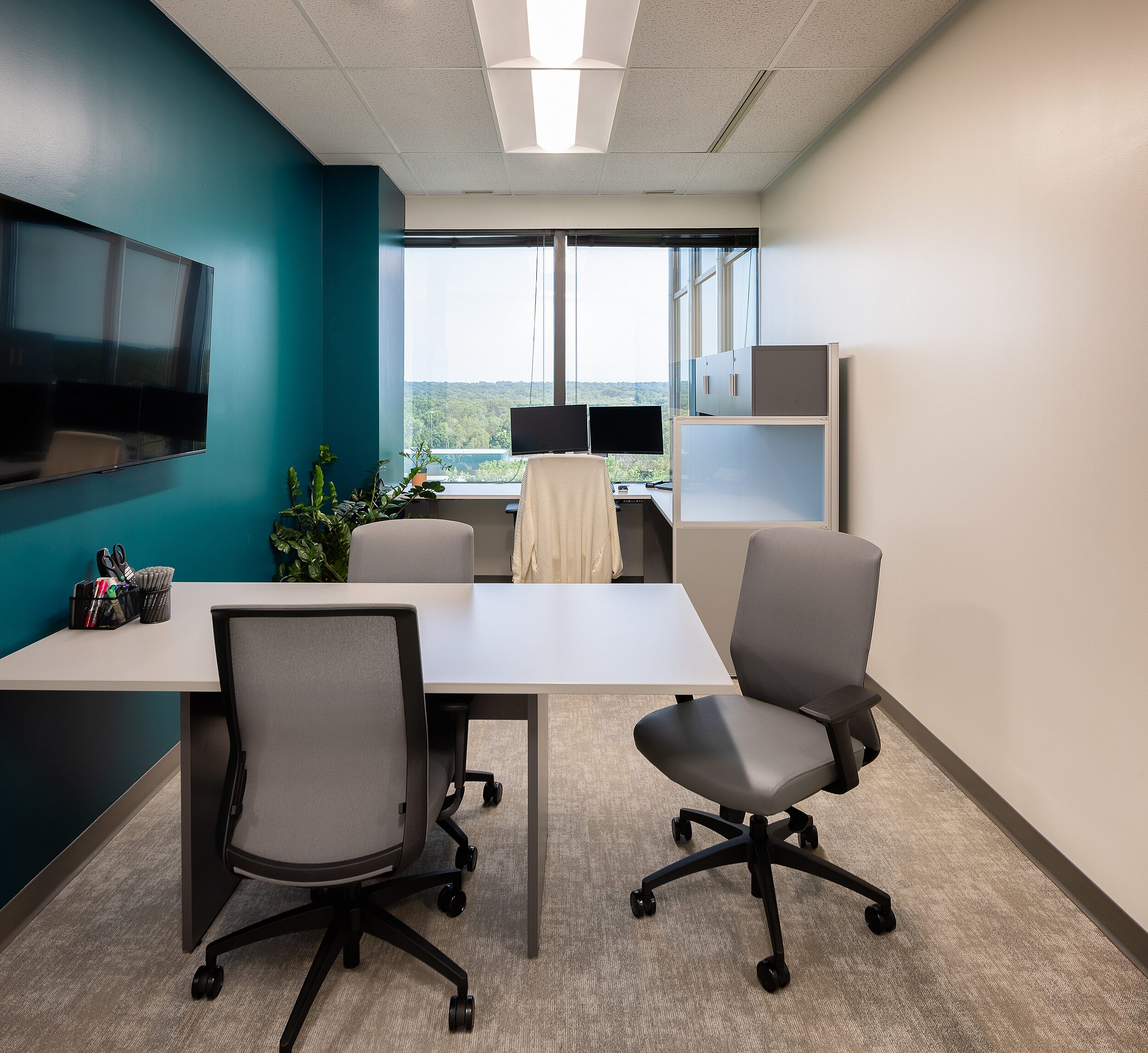 Admin workstation with teal wall and window view. Behind it is a huddle area with a table, three grey rolling chairs and a mounted tv screen