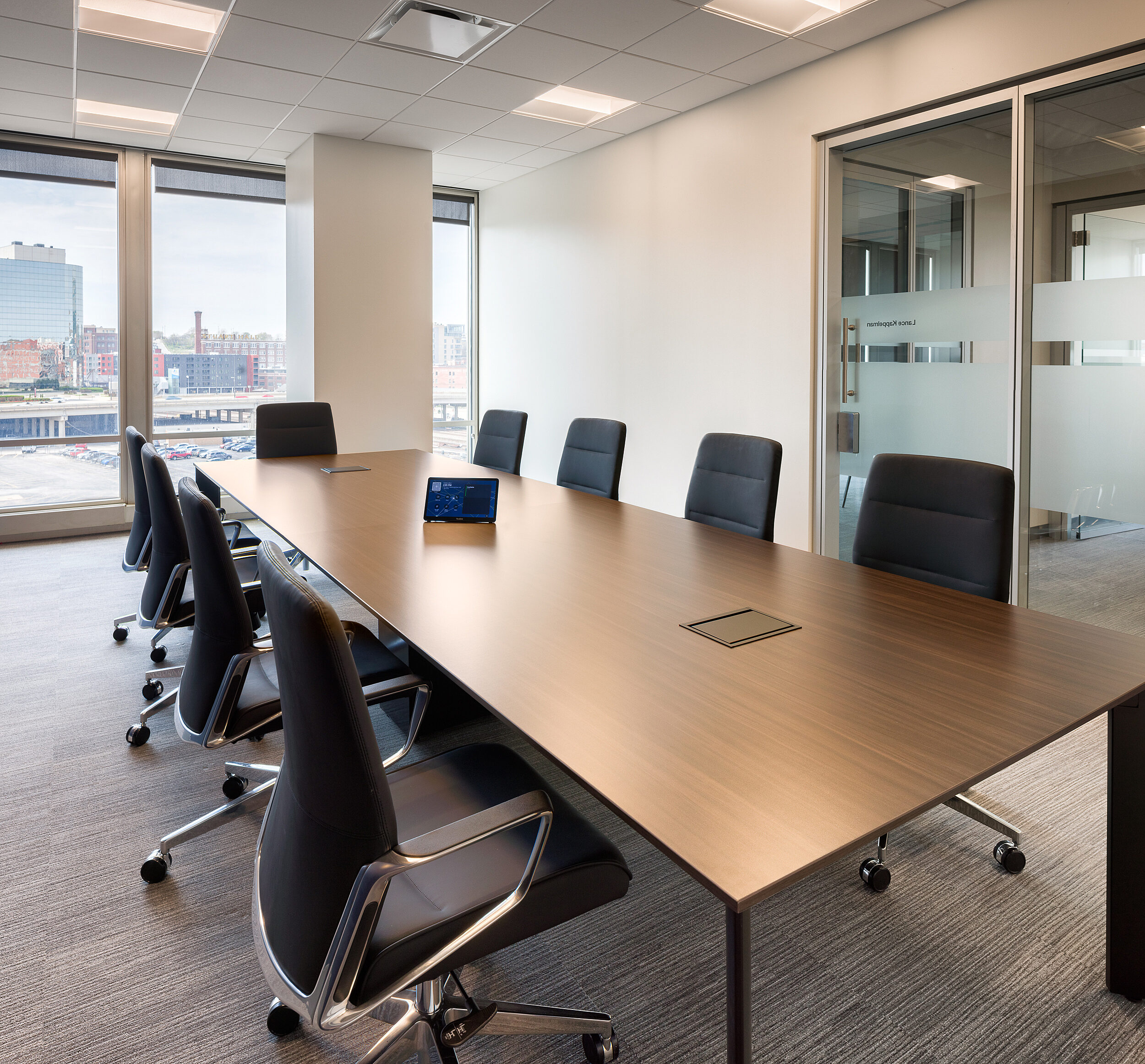 Conference room with a rectangle table and 9 high-backed black leather/chrome chairs. Glass window and door to right side. Window with skyline view.