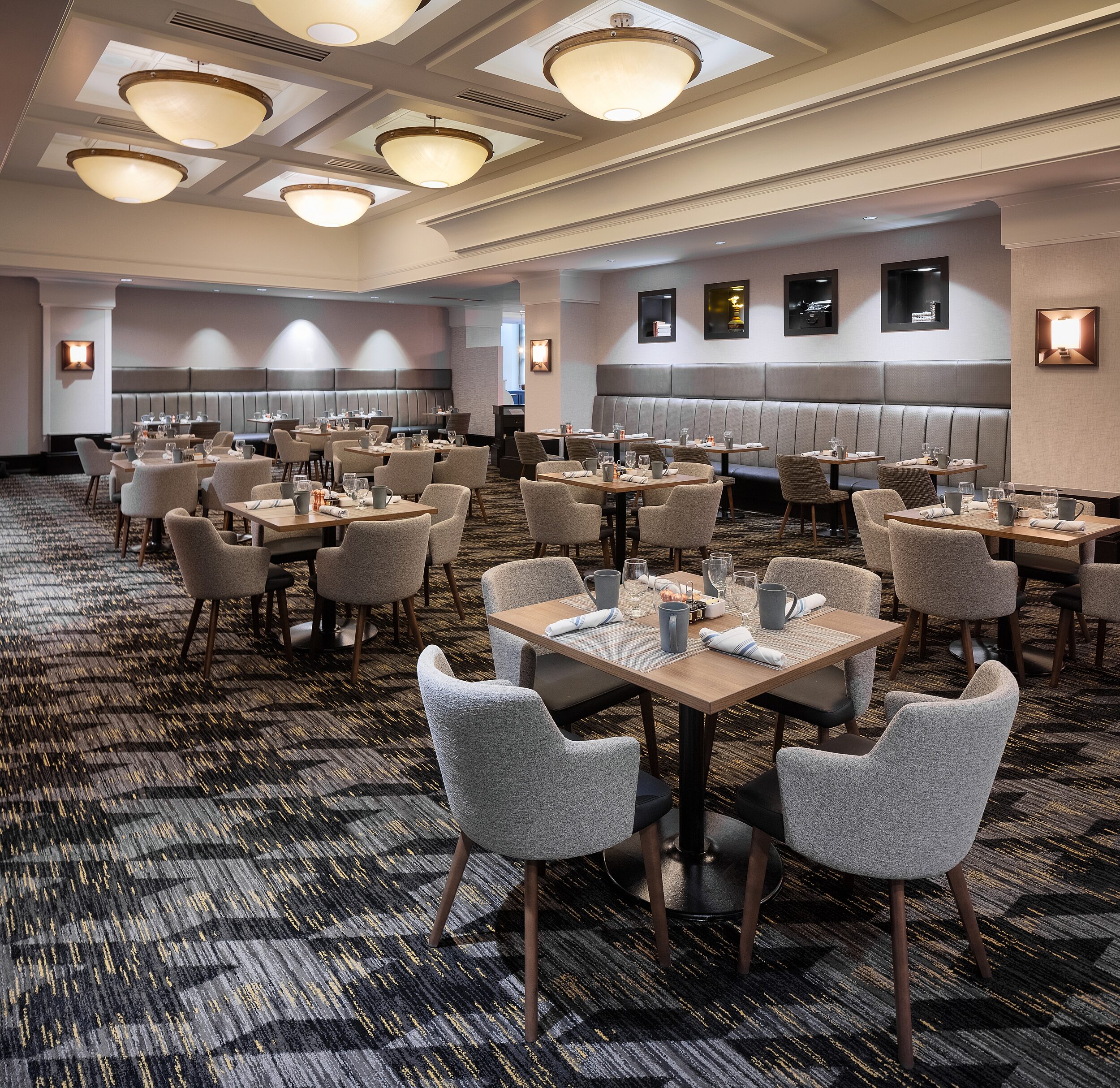 Hotel dining room. Dim lighting. Wood dining tables with mid-back upholstered grey chairs and wooden legs. Banquette seating line all walls. Bold carpet.
