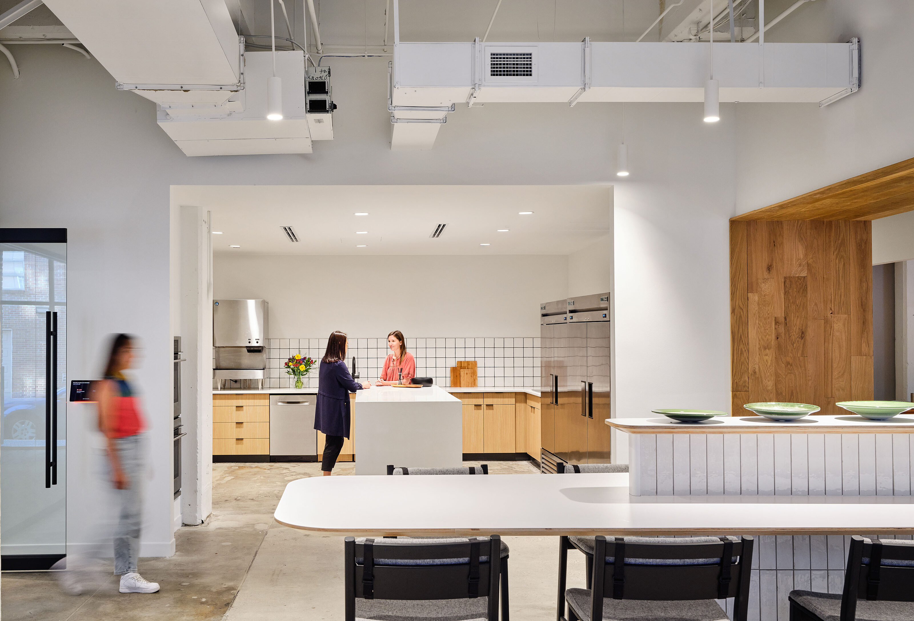 Client's Kitchen/breakroom view with white tile and light wooden cabinets. Foreground is a long counter seating area with tall black stools with backs.
