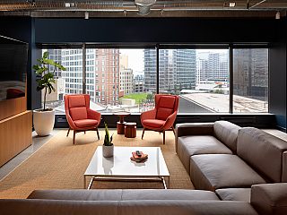 PayIt lounge with view of city buildings. Two orange high-back lounge chairs, a tv, and L-shaped leather sofa, coffee table and industrial ceiling.