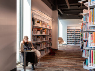 Young adult section of library with shelves of books. A young woman sits in a chair with a small table and reads a book in front of window. Moody lighting.