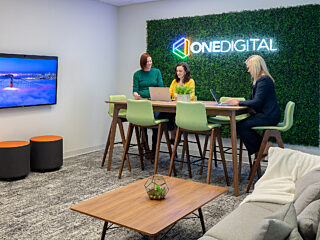 3 women working at a counter-height wood table in  a room with a couch/coffee table. They are seated in front of a "moss" wall with a neon OneDigital logo