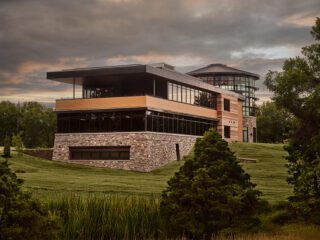 Moody photo of side view of building with multiple floors of rock, black glass, and wood. Tall rock tower with glass with black roof. Architectural design