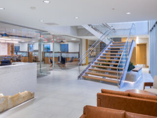 1st floor reception area with modern wood/glass staircase going up. Glass walls show work area with cubicles and other furnishings. Brown leather couches.