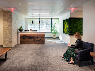 Compass Minerals Reception -tiled wall and moss feature. Receptionist sits and desk and woman in green/black dress and leather jacket sits in a blue chair.