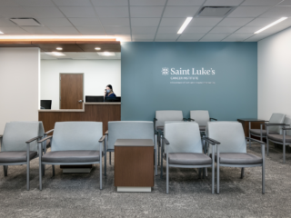 Saint Lukes South - Patient waiting area with blue and grey chairs in front of blue wall with "Saint Luke's Cancer Institute" and reception desk and worker