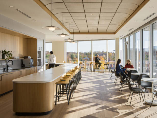 National Advisors Trust breakroom. Two ladies sit at cafe table talking, woman sits at high cafe table working on laptop, a man walks through the kitchen