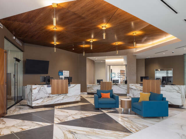 Bank lobby with marble flooring and counters, wood accents on doors, desks and as accent in ceiling. Blue lounge chair seating with pillows and side table