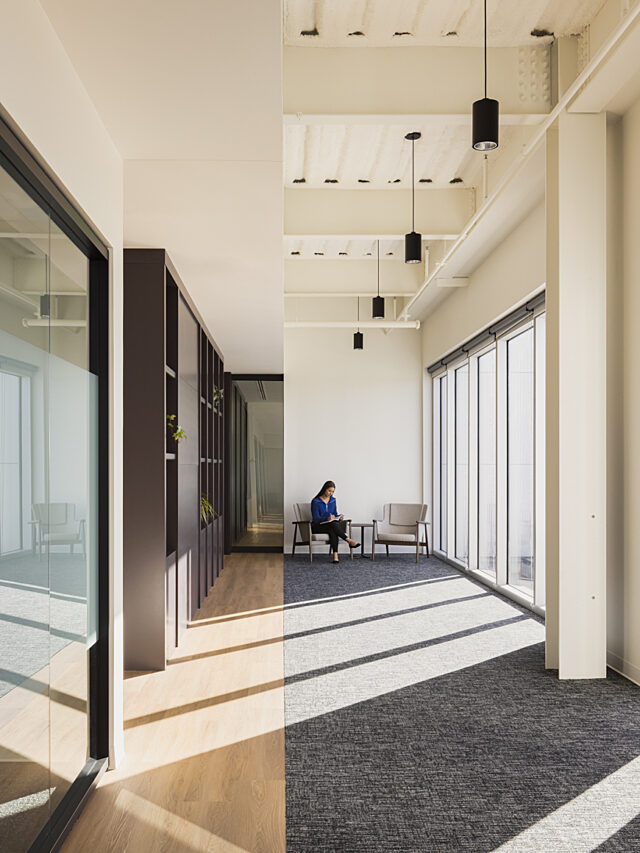 A long windowed hallway with rays of sun coming across it. A girl sits in one of two lounge chairs at very end. White walls and black pendant lighting.
