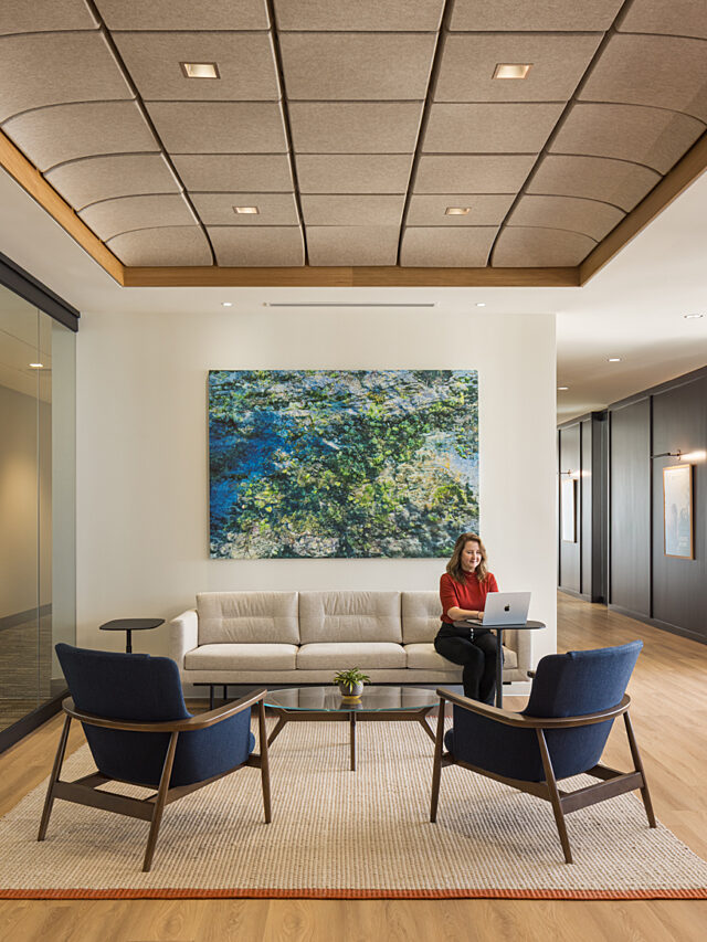 National Advisors Trust lobby with couch under a modern painting. Woman in red works on a laptop computer. Two blue/wood lounge chairs across from her.