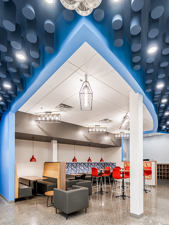 Vertical slide of Schwan's Co's breakroom with industrial lighting, a blue ceiling feature, and various types of seating in red, blue, grey, and yellow.