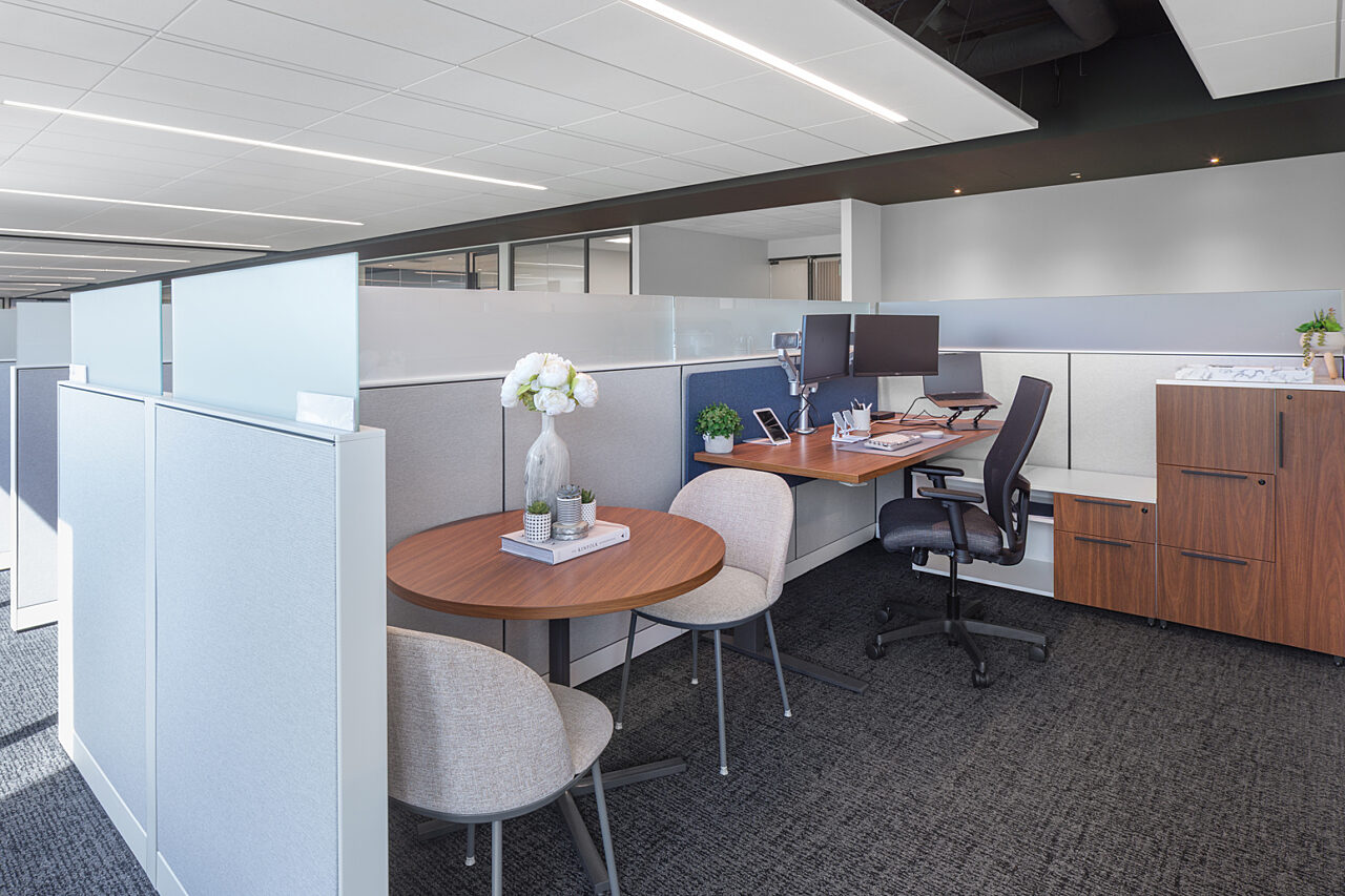 Manager cubicle workstation with height adjustable desks with side cabinet and locker. A small round conference table with 2 chairs sits next to desk.