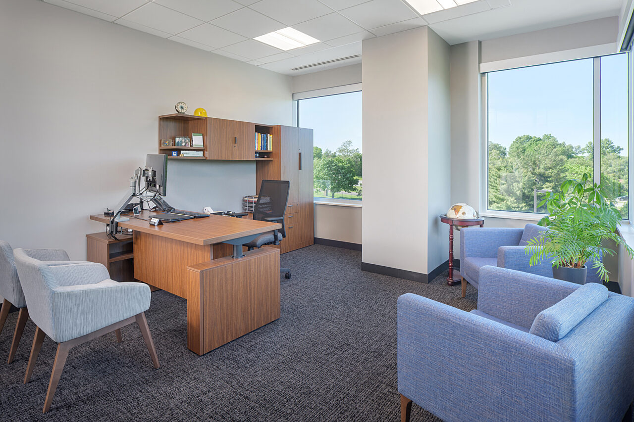 Executive Office with wooden height adjustable desk with upper cabinets and storage locker. Two off-white side chairs and 2 blue lounge chairs by windows