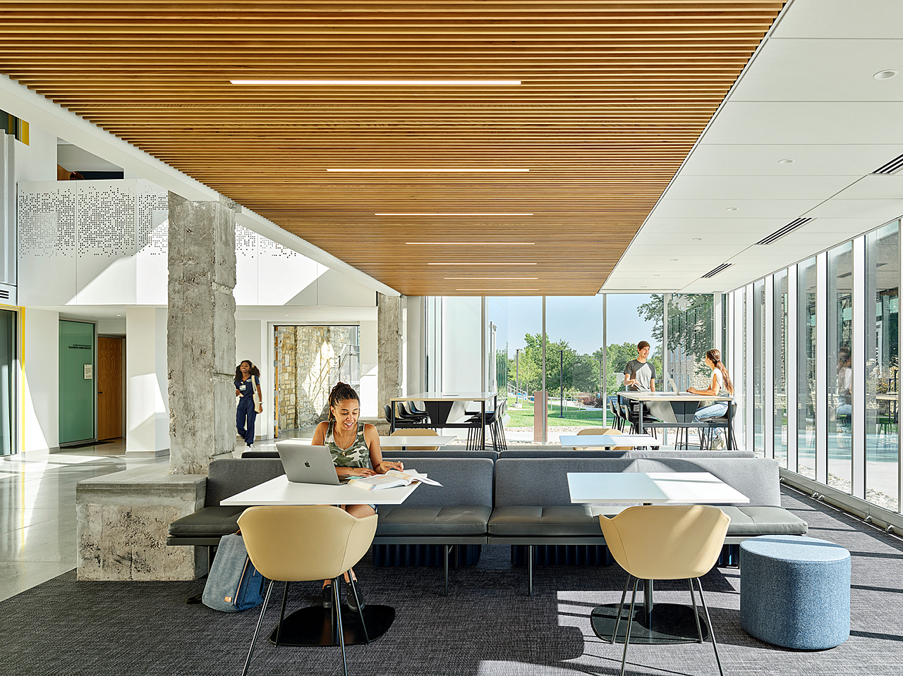 Rockhurst University open study area with students studying at high-top tables and bench seating areas. Wooden slat ceiling and windows line outside walls.