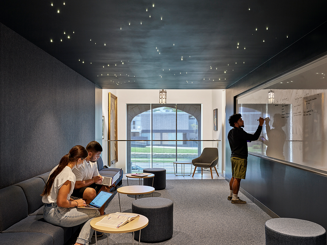 Rockhurst University- Small study room in dark blues with ceiling pin lights resembling starry sky. A man uses a whiteboard. Two others study on benches